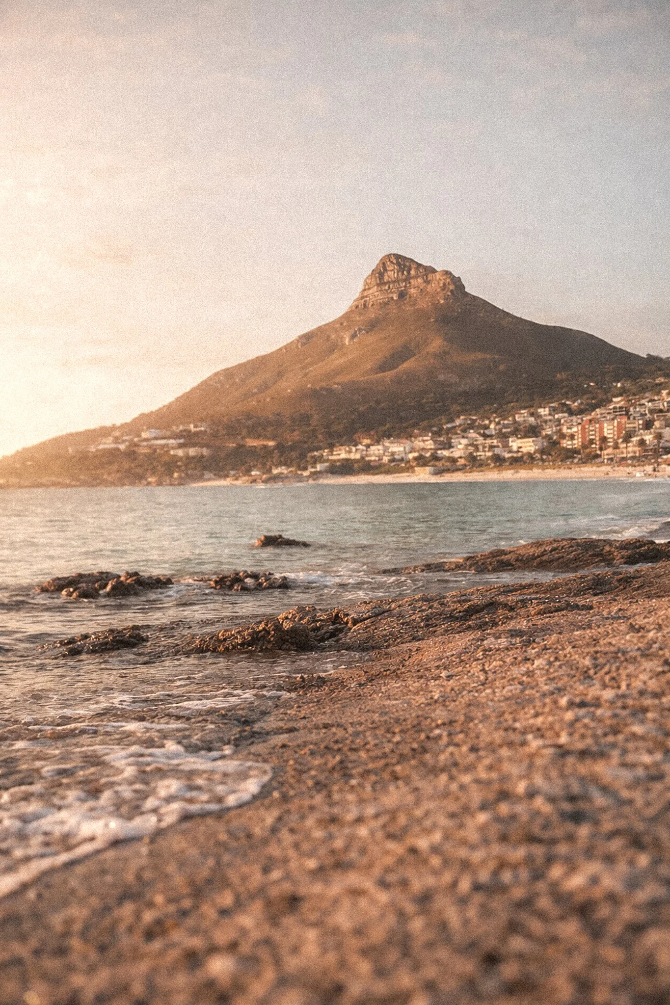 A scenic view of lions head in Cape Town, with rocks and gentle waves in the foreground and a row of houses on the hillside. The scene appears to be during sunset or sunrise.