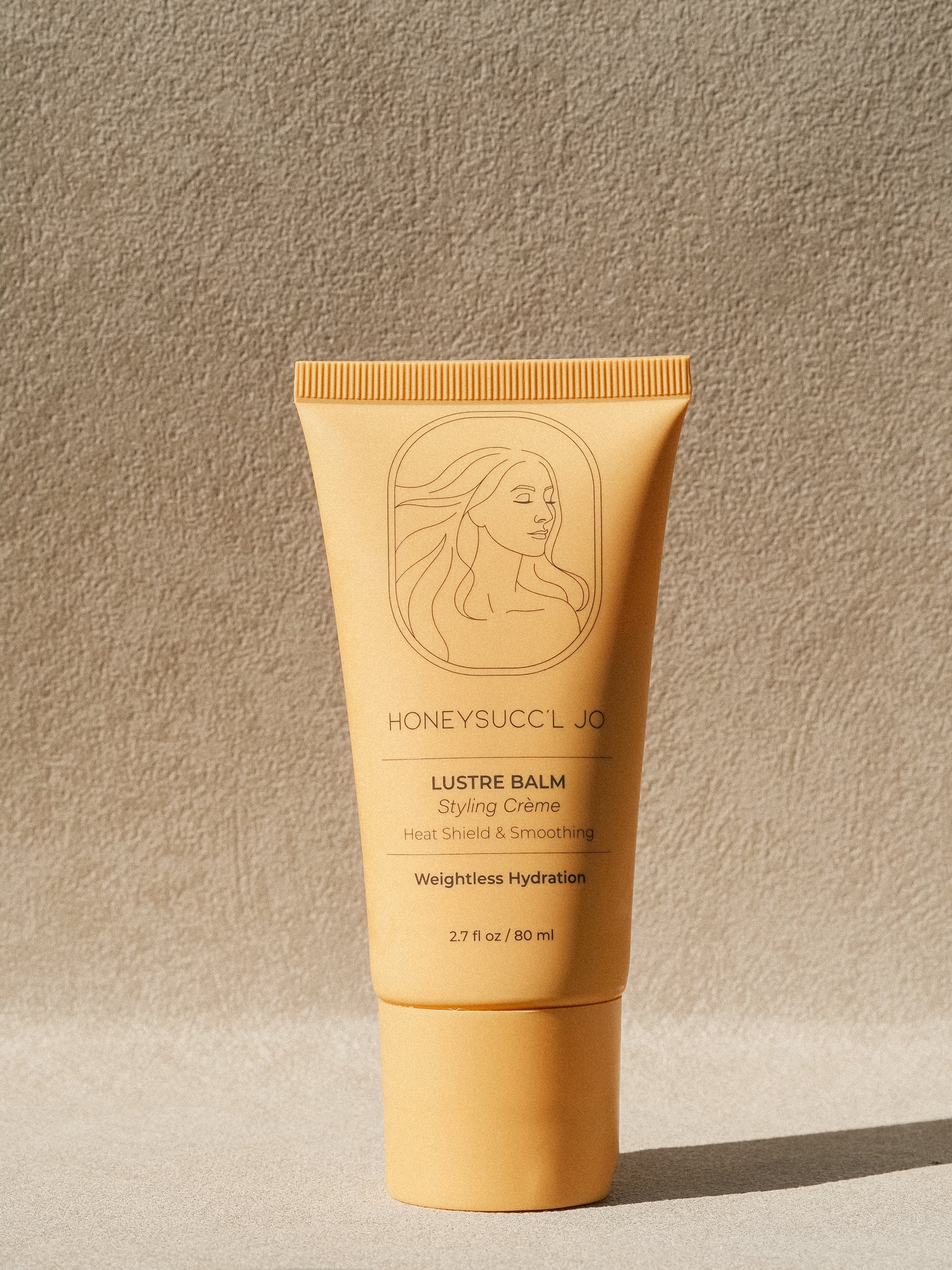 A beige tube of HONEYSUCC'L JO Lustre Balm Styling Crème standing upright on a beige surface with a textured beige background.