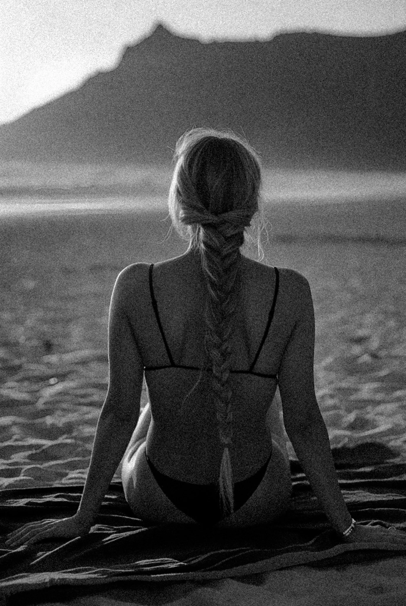 A woman sitting on a beach at sunset, facing away from the camera, with her hair in a braid. She is wearing a bikini and looking toward the ocean and distant mountains.