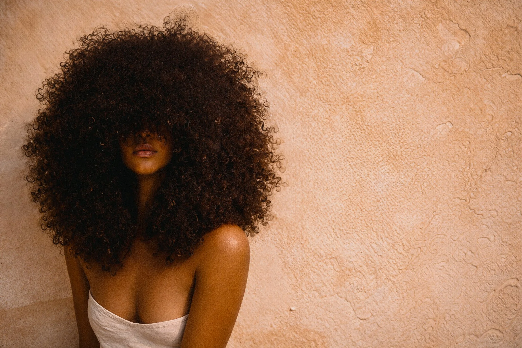 A woman with large, curly hair covering her face, positioned against a textured beige wall.