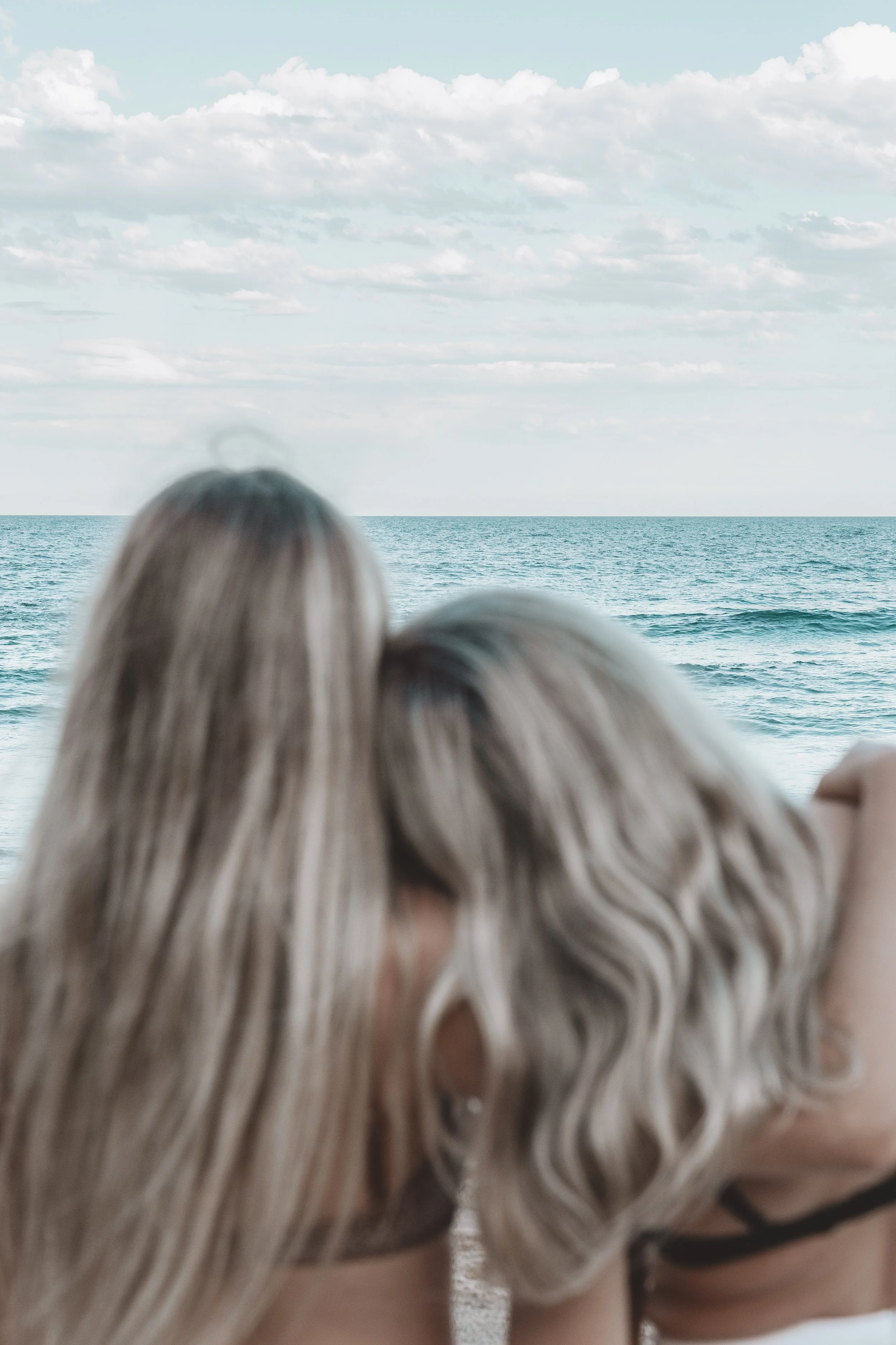 Two women with blonde hair are seen from behind, sitting close together at the beach, looking out at the ocean with a partly cloudy sky.