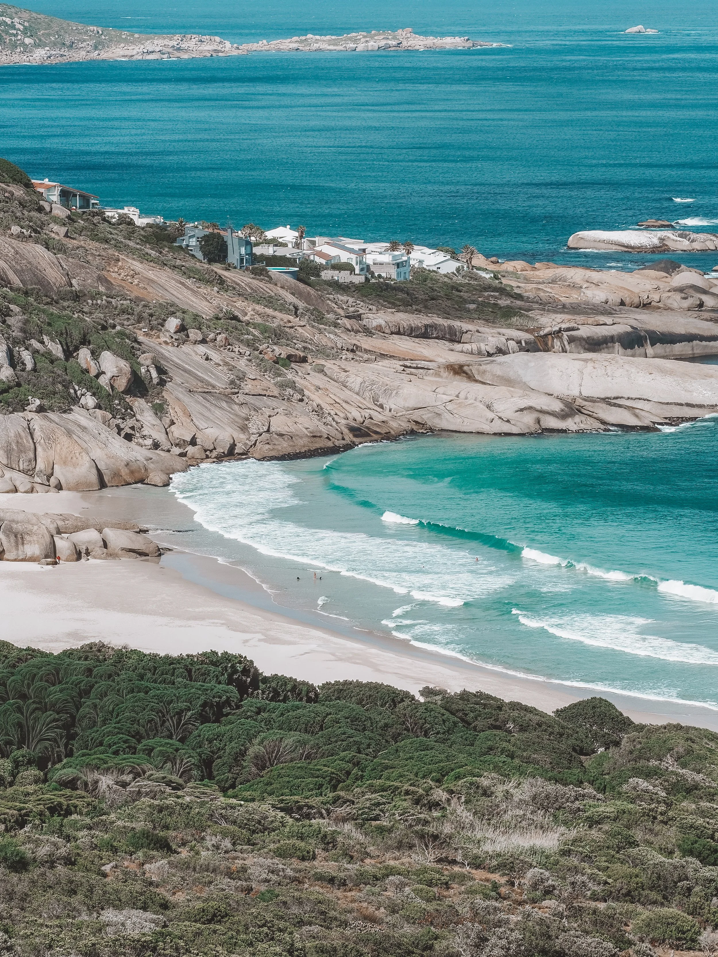 Coastal view with rocky hills, white sandy beach, turquoise ocean waves, and modern houses overlooking the water. Cape Town, haircare line, nature inspired