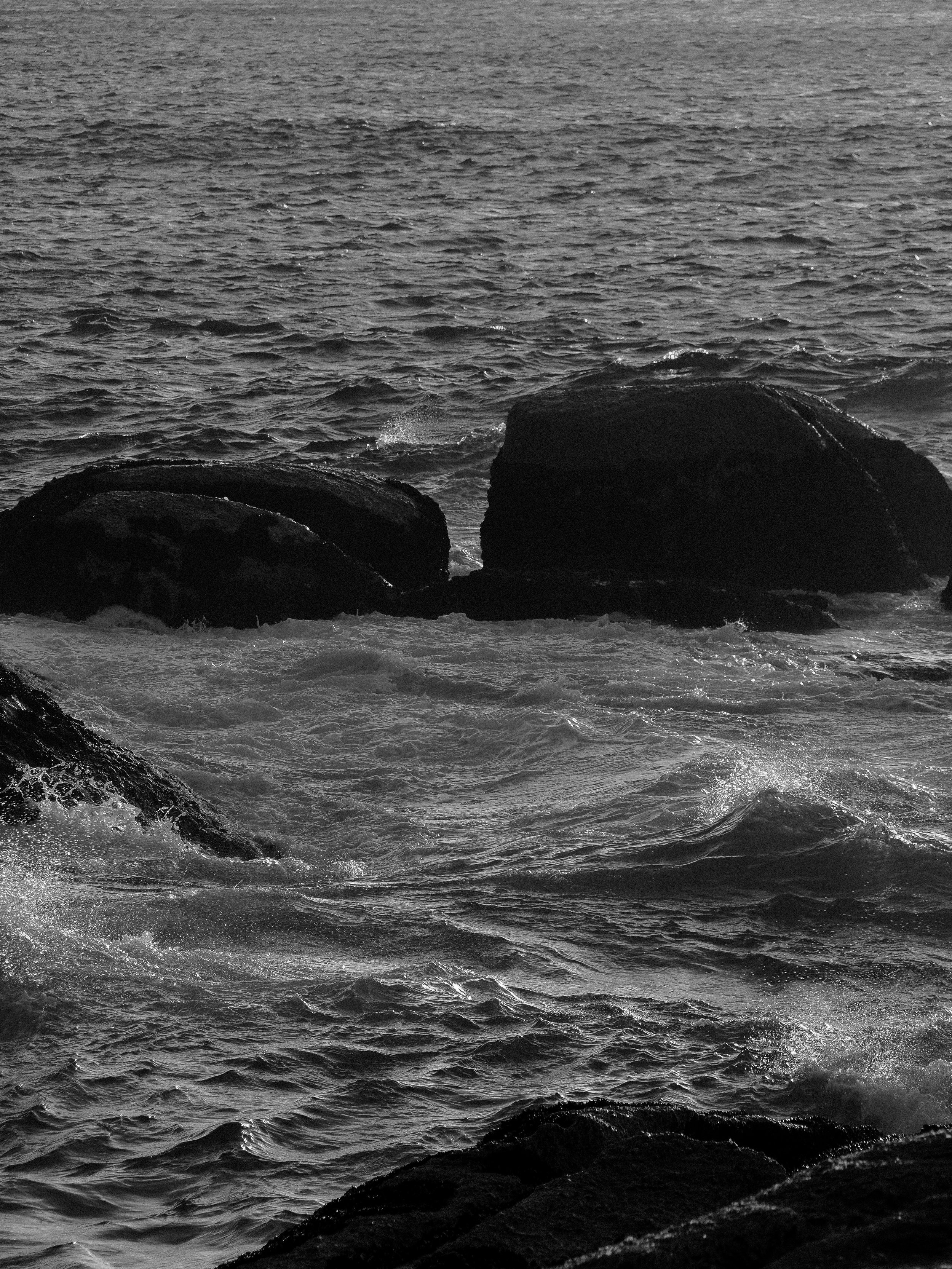 Black and white photo of ocean waves crashing against rocks at the shoreline.