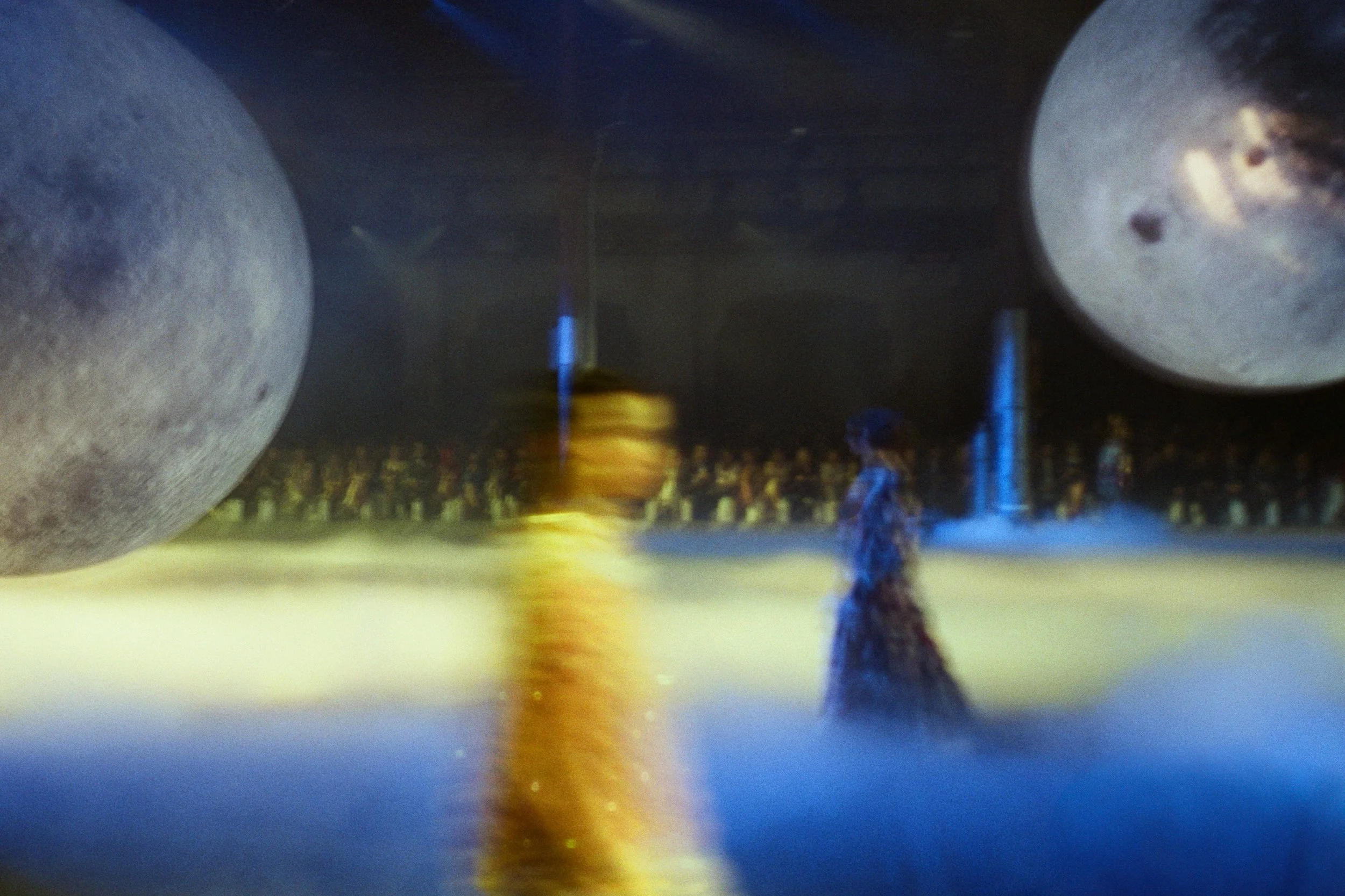 Artistic scene with two people walking in a foggy, surreal environment featuring two large moons in the sky and a fountain in the background.