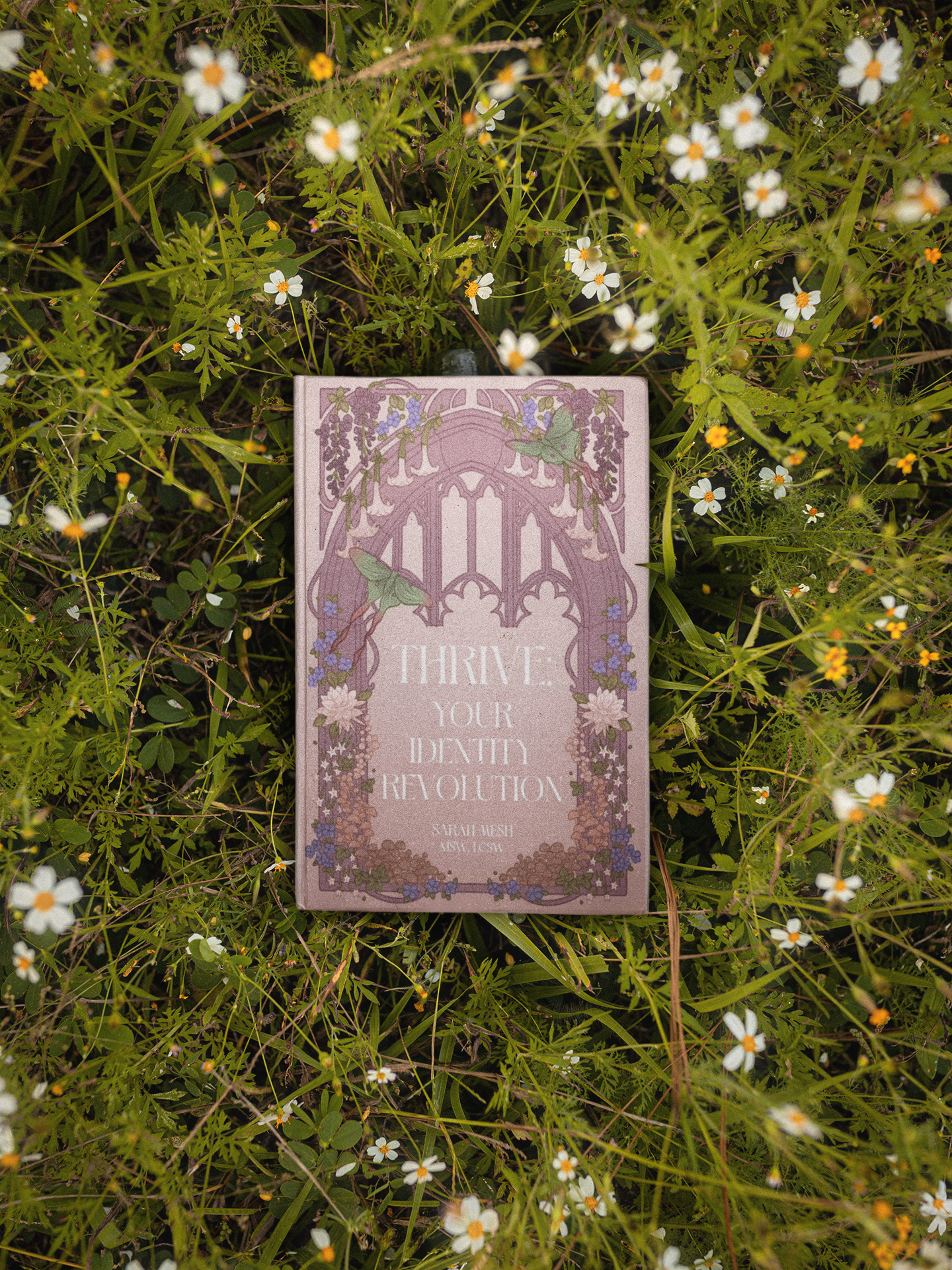 A book titled 'Thrive Your Death Revolution' by Sariah Kuishi resting among small white flowers and green grass.