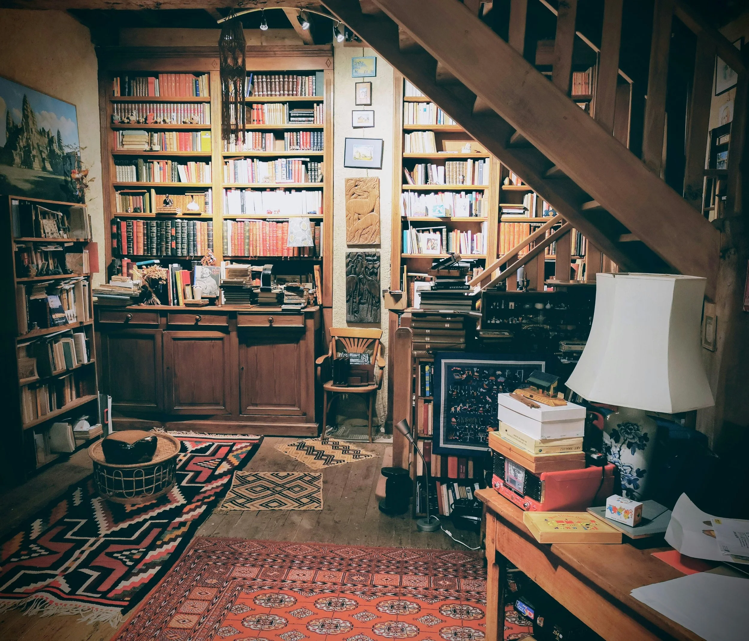 A cozy room with wooden bookshelves filled with books, a staircase leading upstairs, a colorful rug on the floor, and various decorations including framed pictures and artwork.