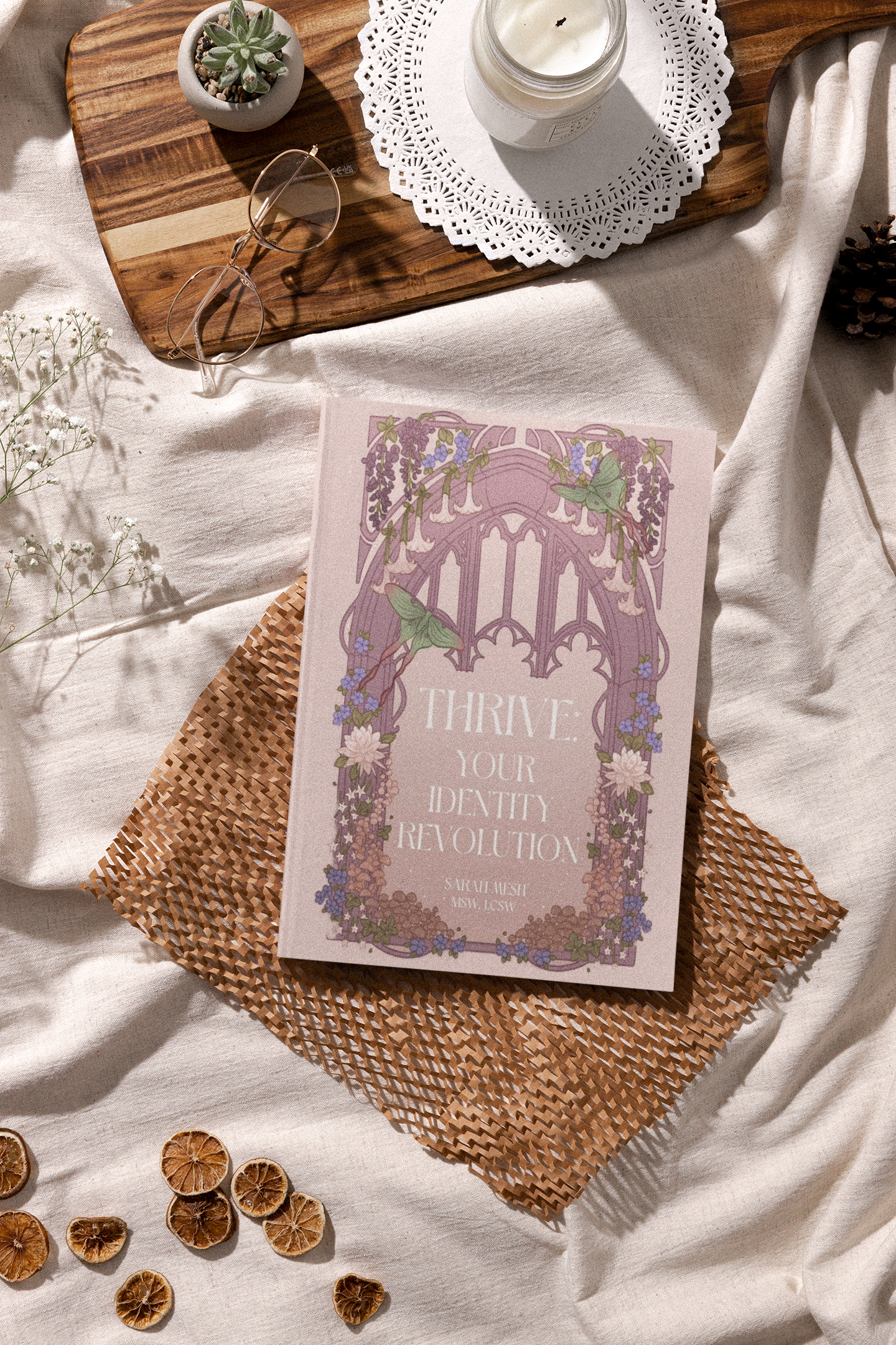 A light pink booklet titled "Thrive Your Identity Revolution" resting on a woven mat, with dried orange slices surrounding it. A wooden tray with a potted succulent, a lit candle in a glass jar on a paper doily, and a pair of glasses are also on the tray, all placed on a white fabric surface.