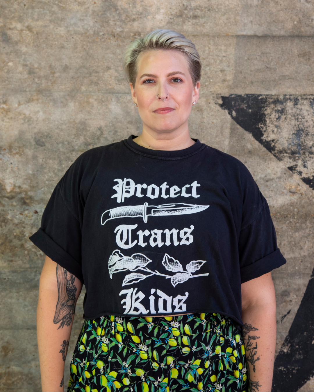 A woman with short, platinum blonde hair standing in front of a textured wall. She is wearing a black t-shirt with white text and graphics that say 'Protect Trans Rights' and features a knife and a branch with leaves, along with a skirt with a lemon and lemon leaf pattern.