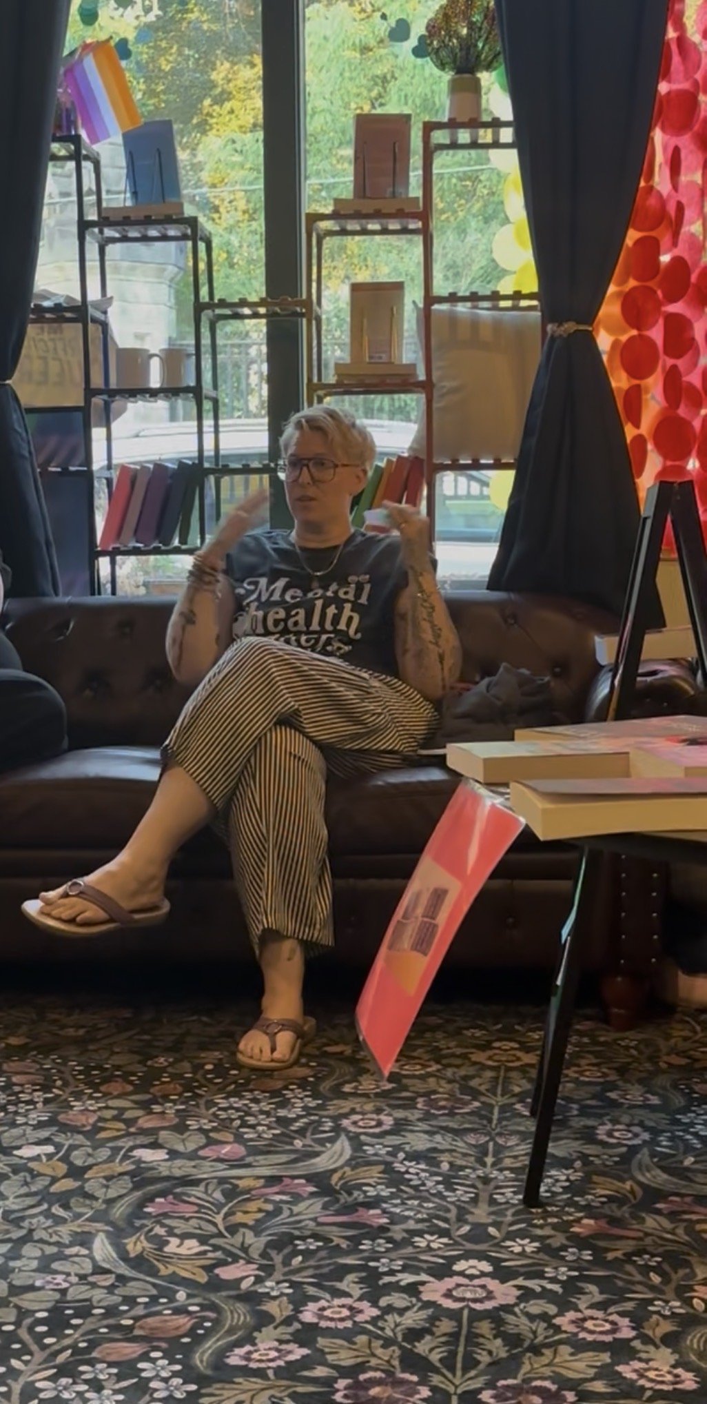 A person with short blonde hair, glasses, and tattoos sitting on a brown leather couch in a bookstore or library. They are wearing a dark t-shirt with white text, striped pants, and flip-flops. The background includes bookshelves, a window with greenery outside, and colorful curtains.