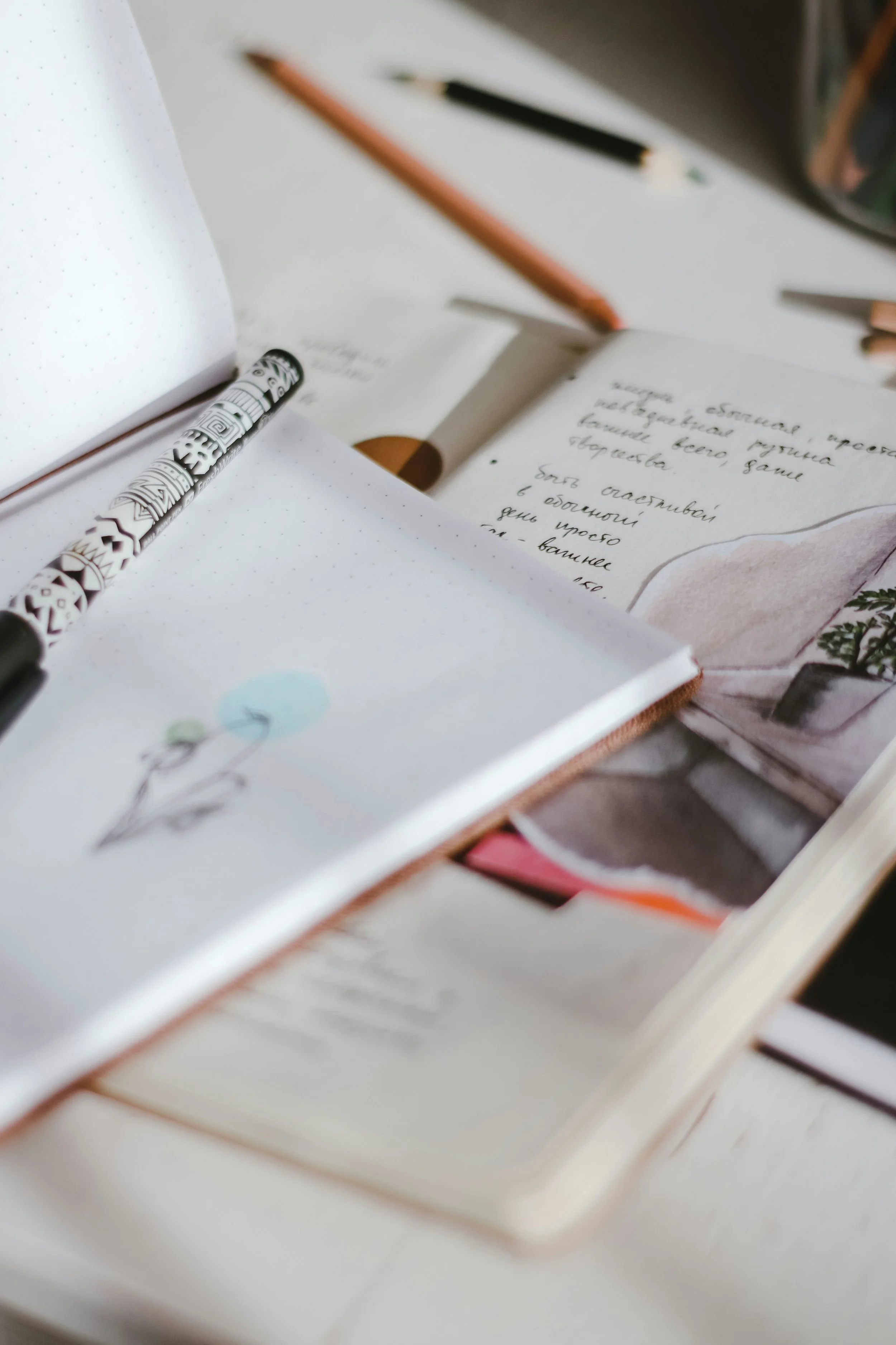 Open notebook with handwritten notes, a decorative pen, a pencil, and a closed book with a floral cover on a white table.