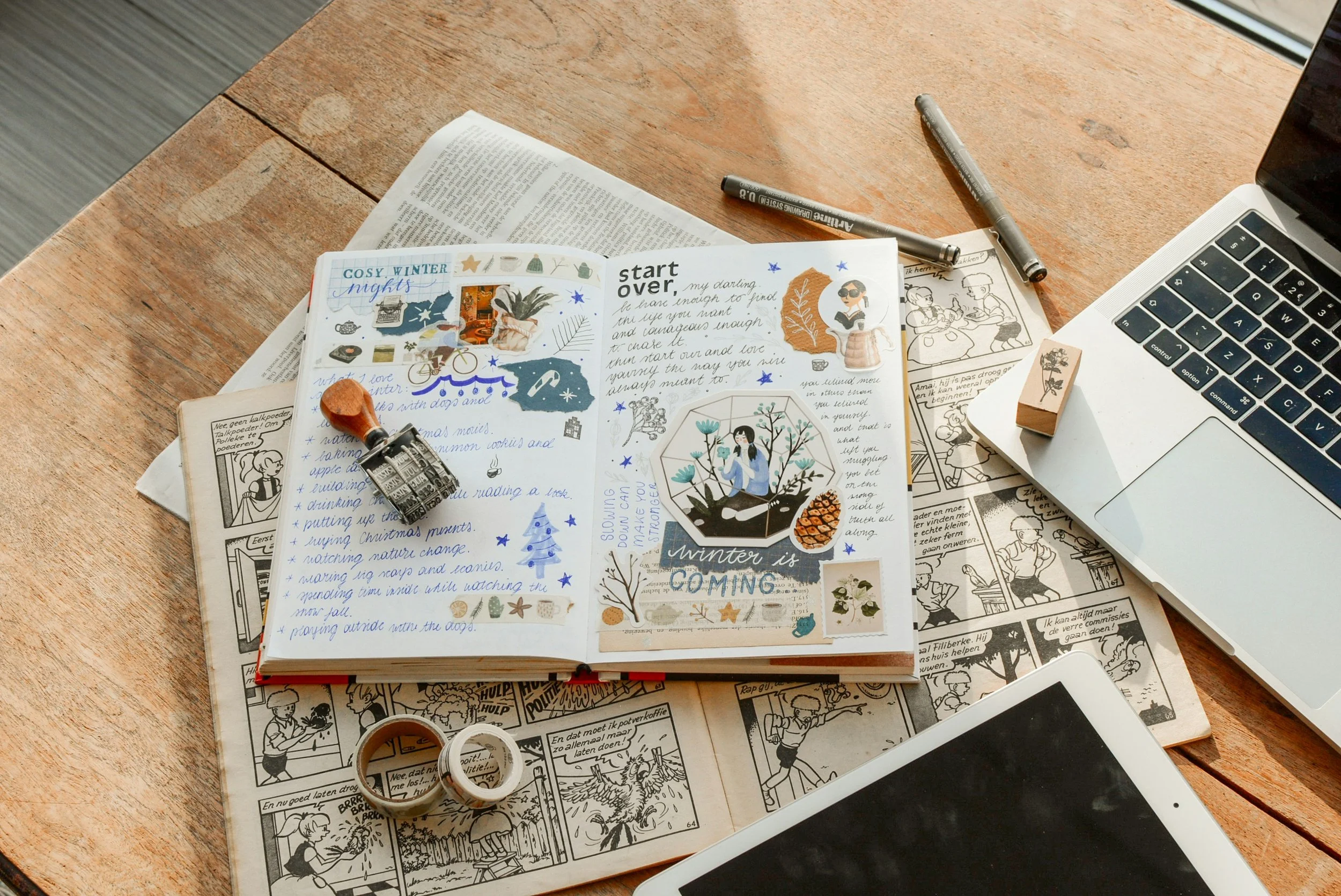 A wooden desk with open books, a journal with colorful stickers and handwritten notes, two markers, washi tapes, a paper clip, a laptop, and some comic pages.