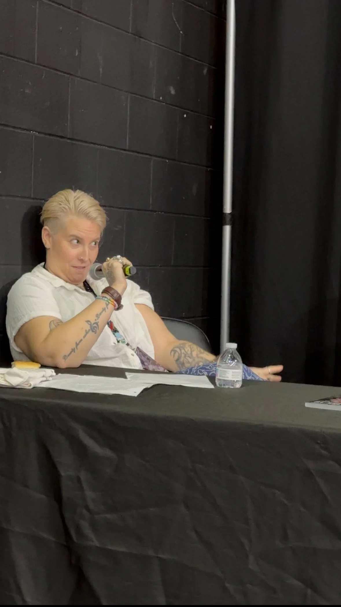 Person with short blonde hair and tattoos sitting at a table, holding a microphone in one hand, with a water bottle and papers on the table, against a black brick wall background.