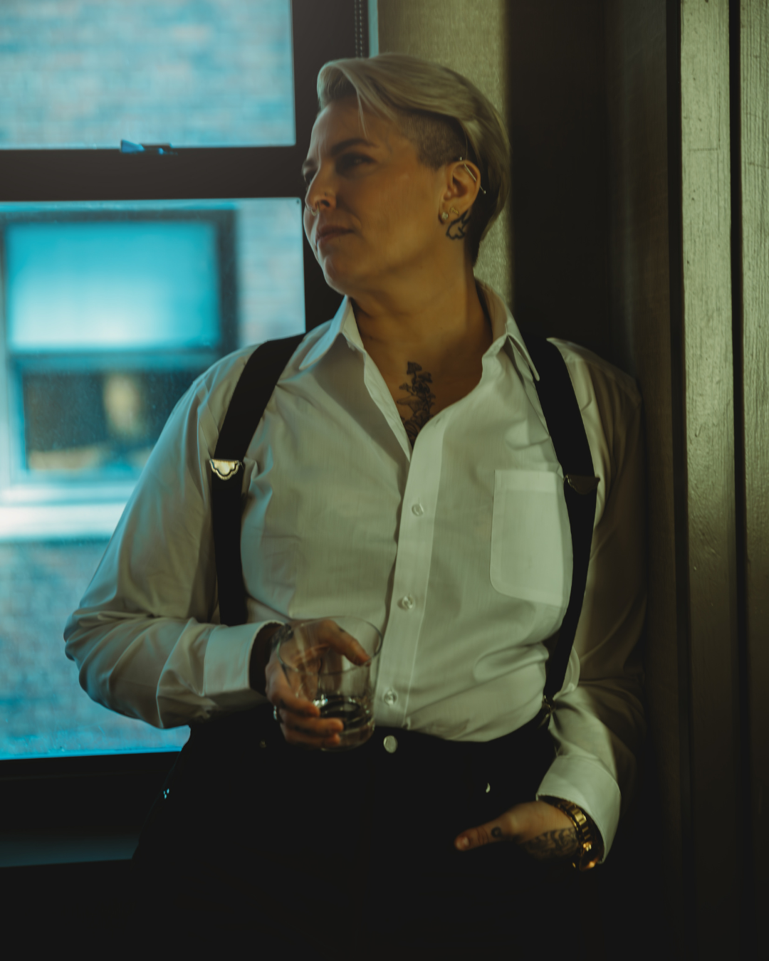 A person with short, styled hair leaning against a wooden wall, wearing a white button-up shirt, suspenders, earrings, tattoos, and holding a glass of drink, inside a room with a window.