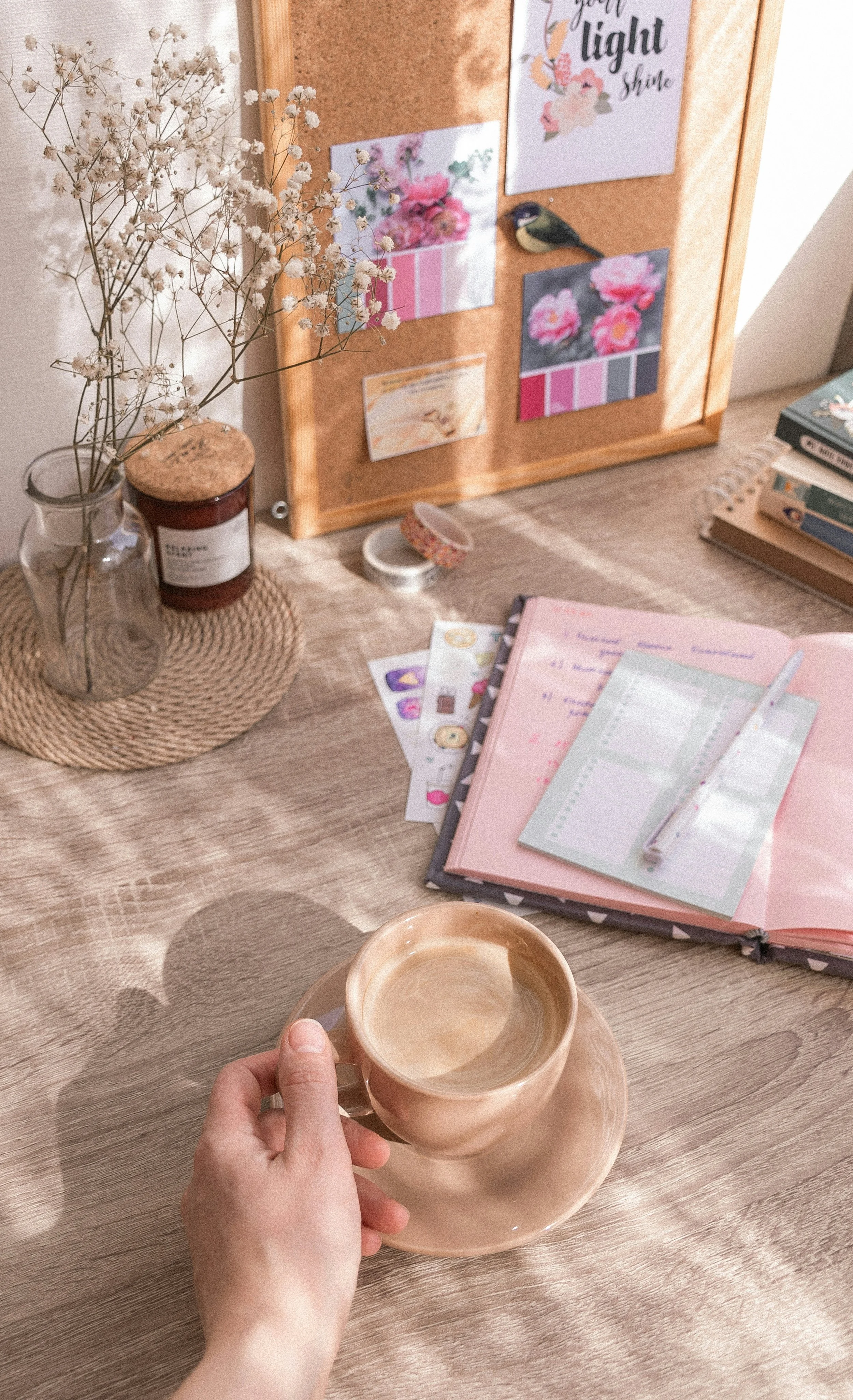 Person holding a ceramic cup of coffee or tea over a wooden table, with a pink notebook, sticky notes, a corkboard with motivational quotes and artwork, a vase with dried flowers, and a candle on the table.