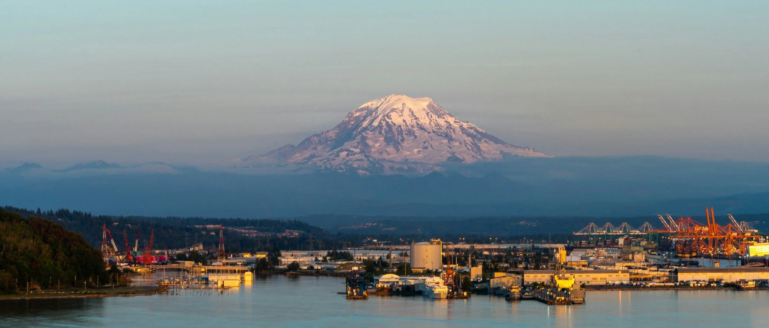 View of Mount Fuji with snow-covered peak, surrounding mountains, and an industrial harbor with cranes in the foreground near a body of water.