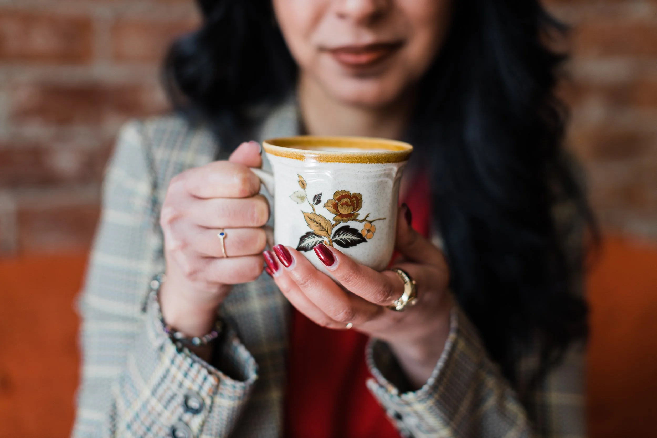 Person with dark hair holding a ceramic mug with floral design, wearing a plaid jacket, rings, and bracelets, against a blurred brick wall background.