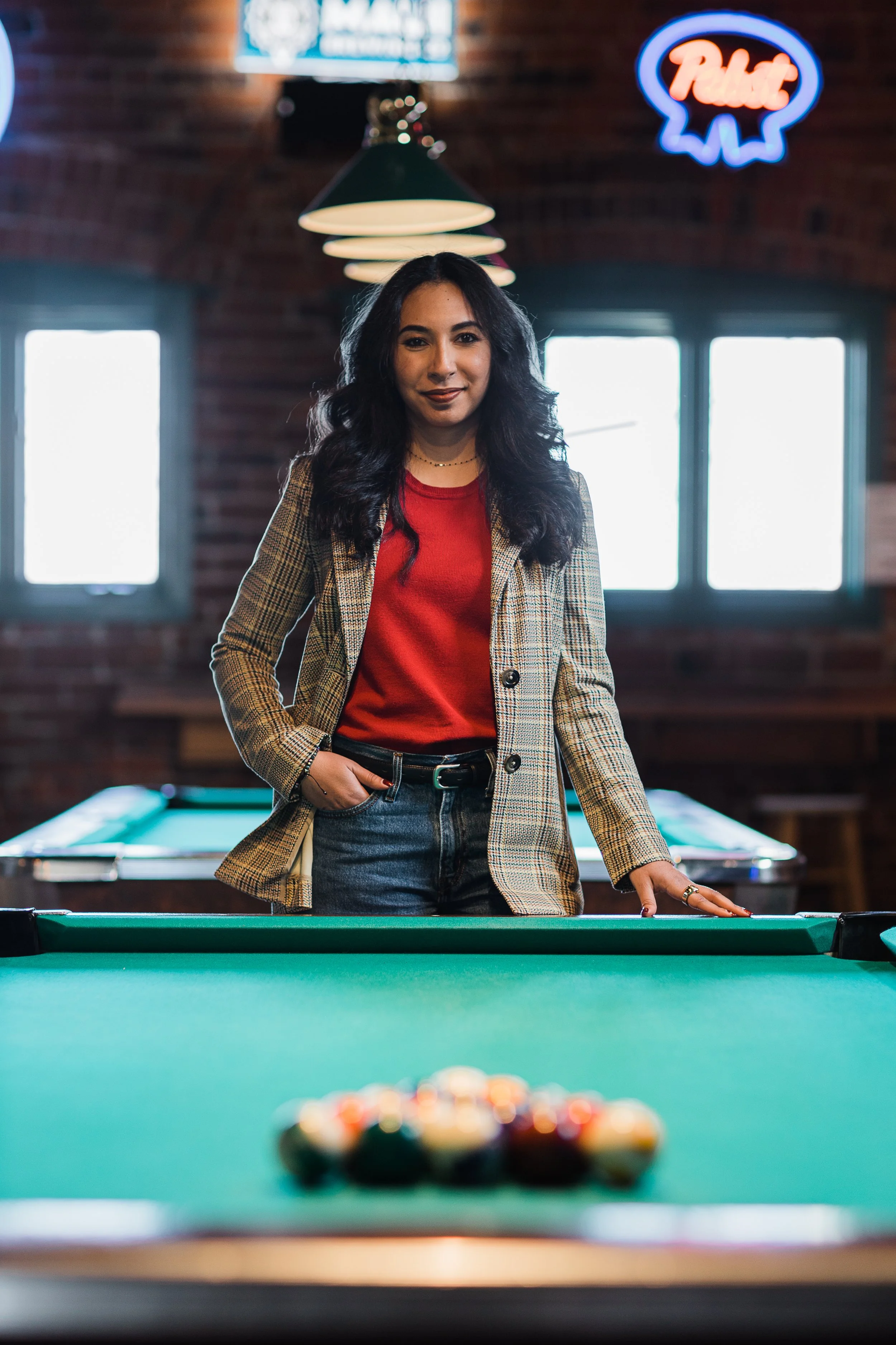 A woman with long dark hair, wearing a plaid blazer and red top, standing at a pool table in a dimly lit, brick-walled bar with neon signs.