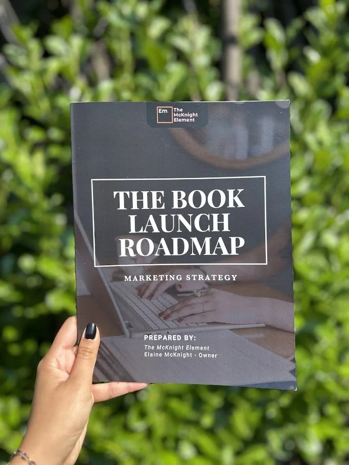 Hand holding a book titled 'The Book Launch Roadmap: Marketing Strategy' by Elaine McKnight, outside with greenery in the background.