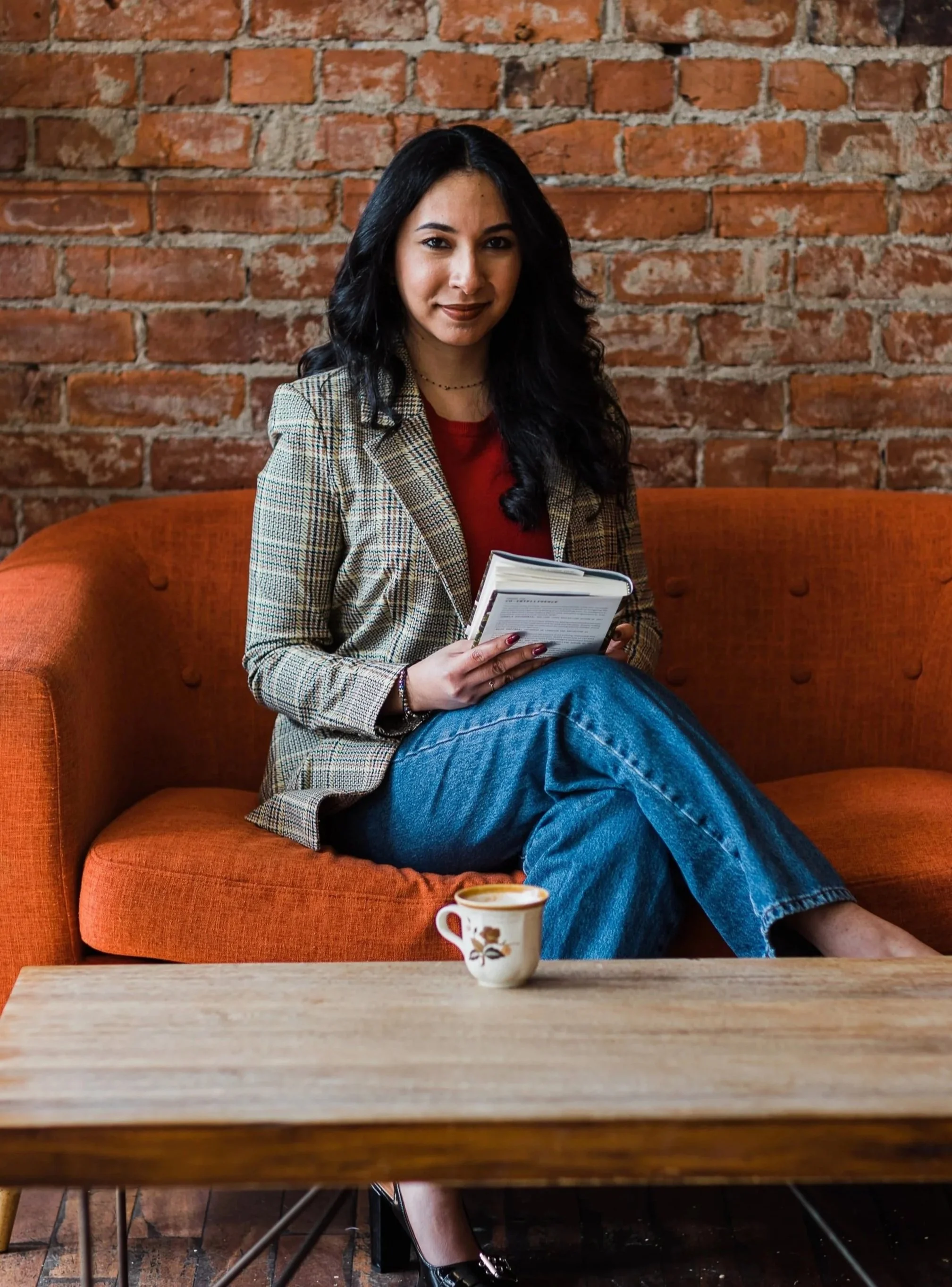 A woman with long black hair, wearing a plaid blazer, red top, and blue jeans, sitting on an orange sofa with a brick wall background, holding a book, with a coffee cup on the table in front.