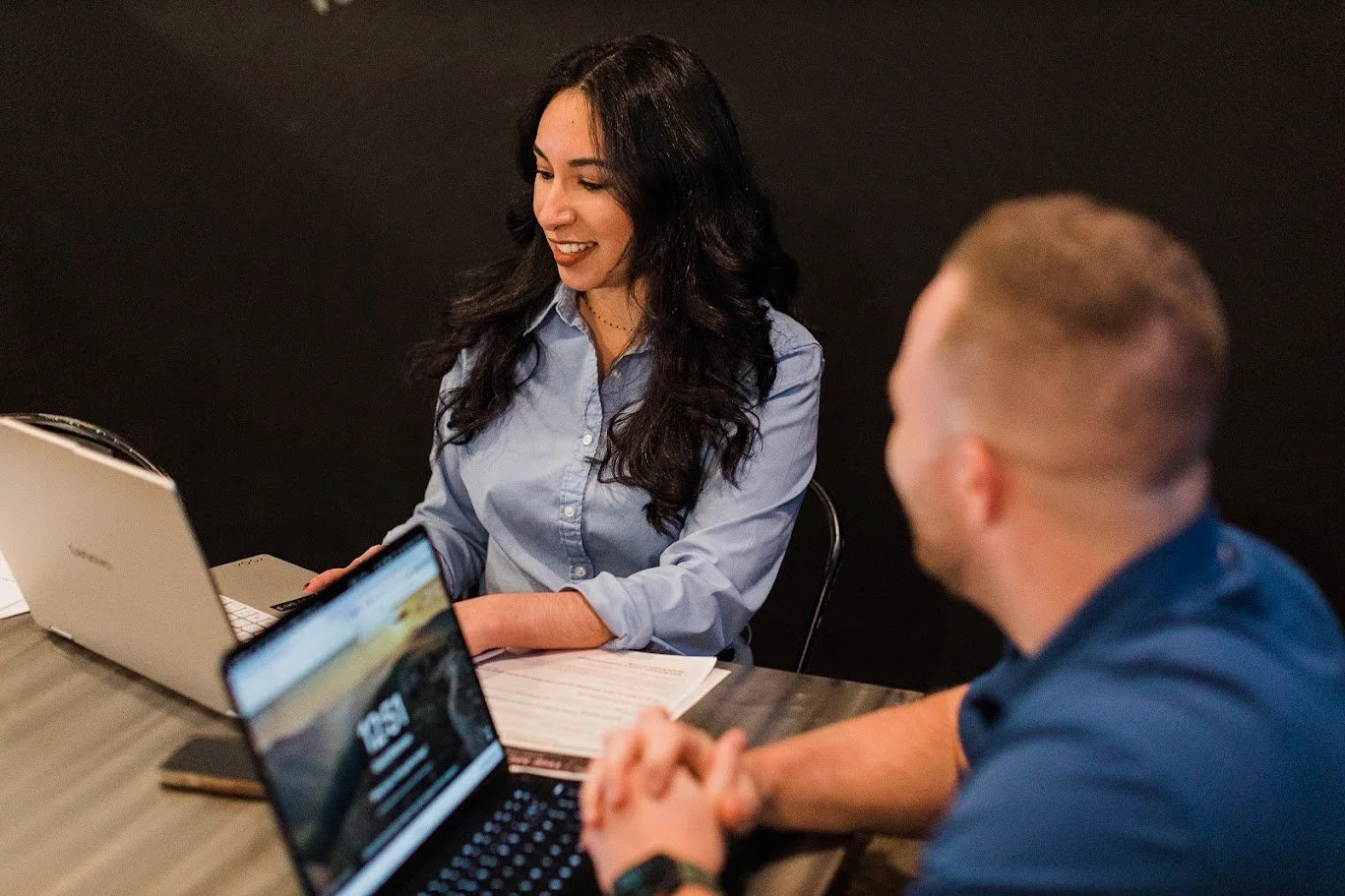 Two people sitting at a table with laptops and notebooks, engaged in a discussion. The woman is smiling and wearing a light blue shirt, and the man has short blond hair, wearing a dark shirt.