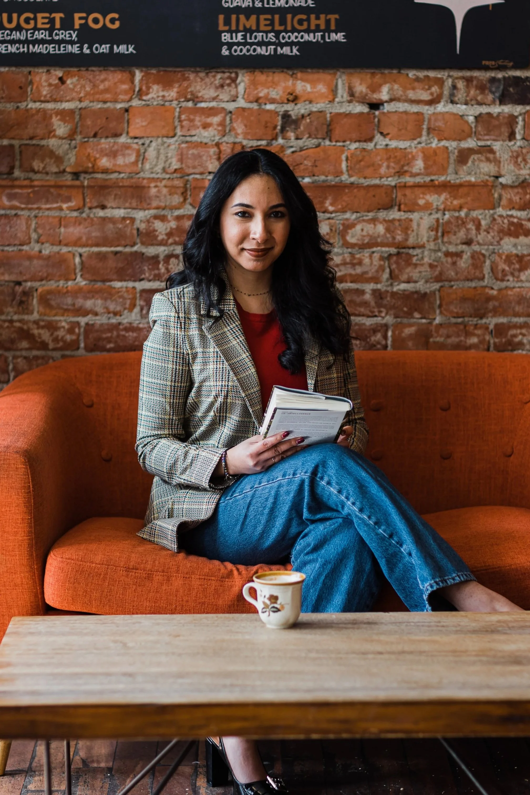 A woman with black wavy hair, wearing a plaid blazer and blue jeans, sitting on an orange sofa with a book in her hands in a cafe with exposed brick walls. A coffee cup with floral design is on the wooden table in front of her.