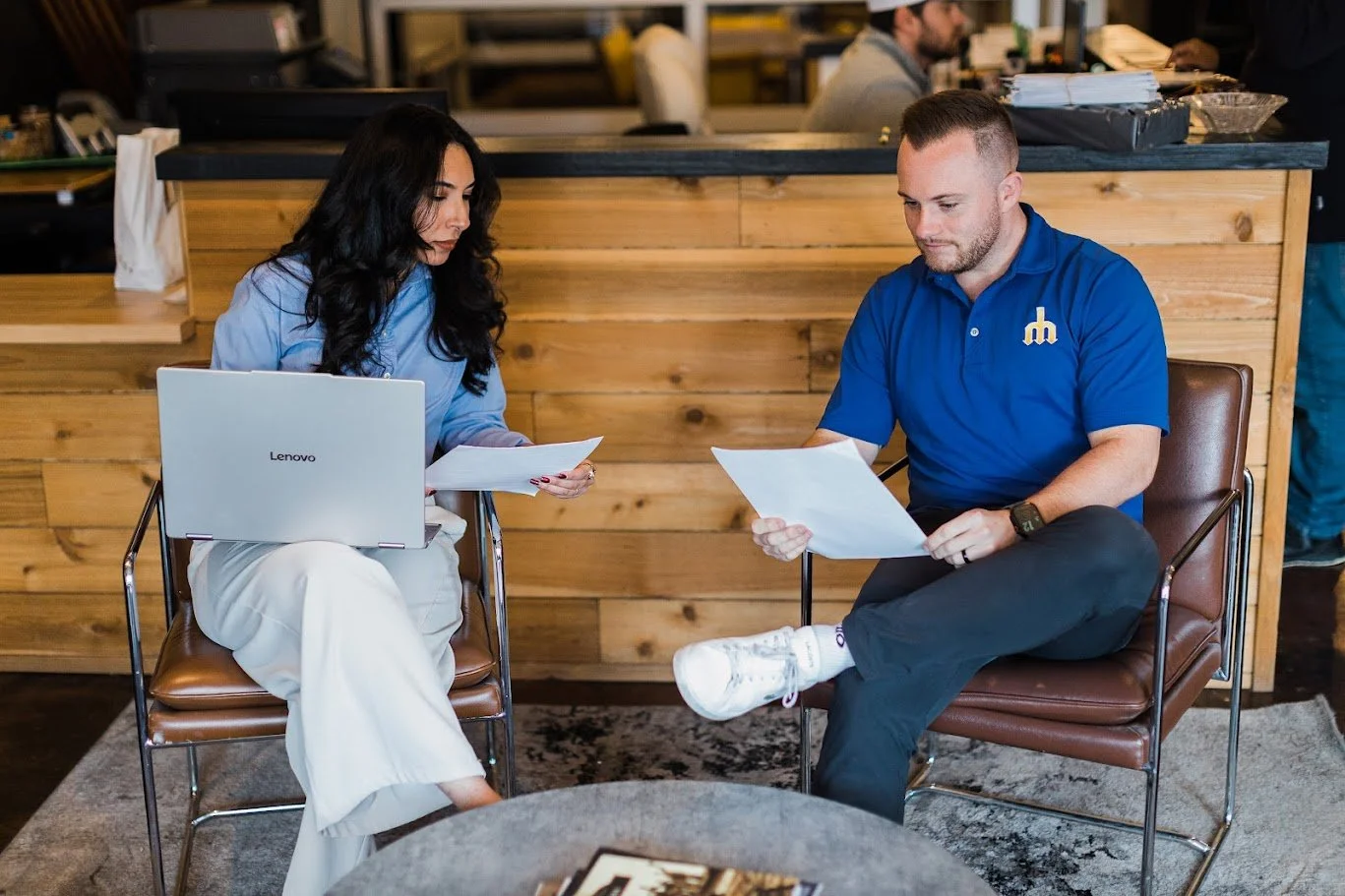 Two people sitting and discussing documents in a cozy, modern office or cafe setting with a wooden counter in the background. One person has dark hair and a laptop, the other has short hair and is wearing a blue polo shirt.