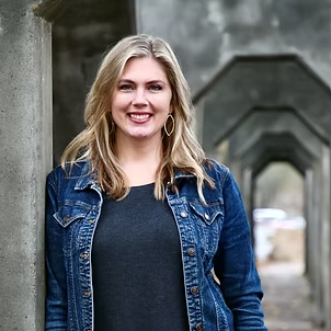 A smiling young woman with blonde hair, wearing a denim jacket over a dark shirt, standing outdoors with a stone archway in the background.