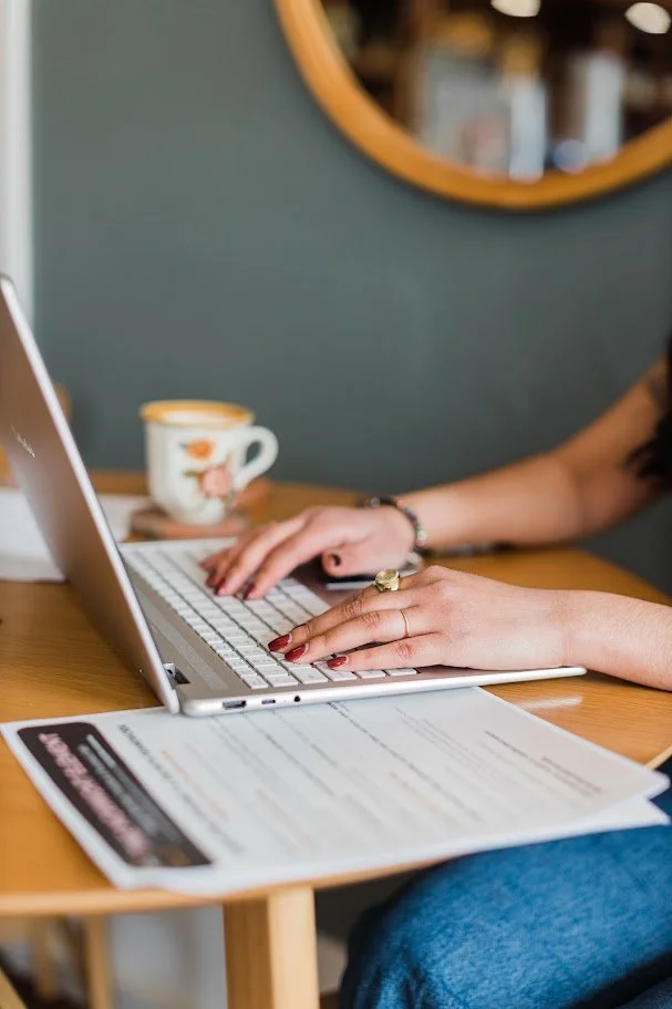 Person typing on a silver laptop at a wooden table, with a ceramic mug in the background and a document in front of the person.