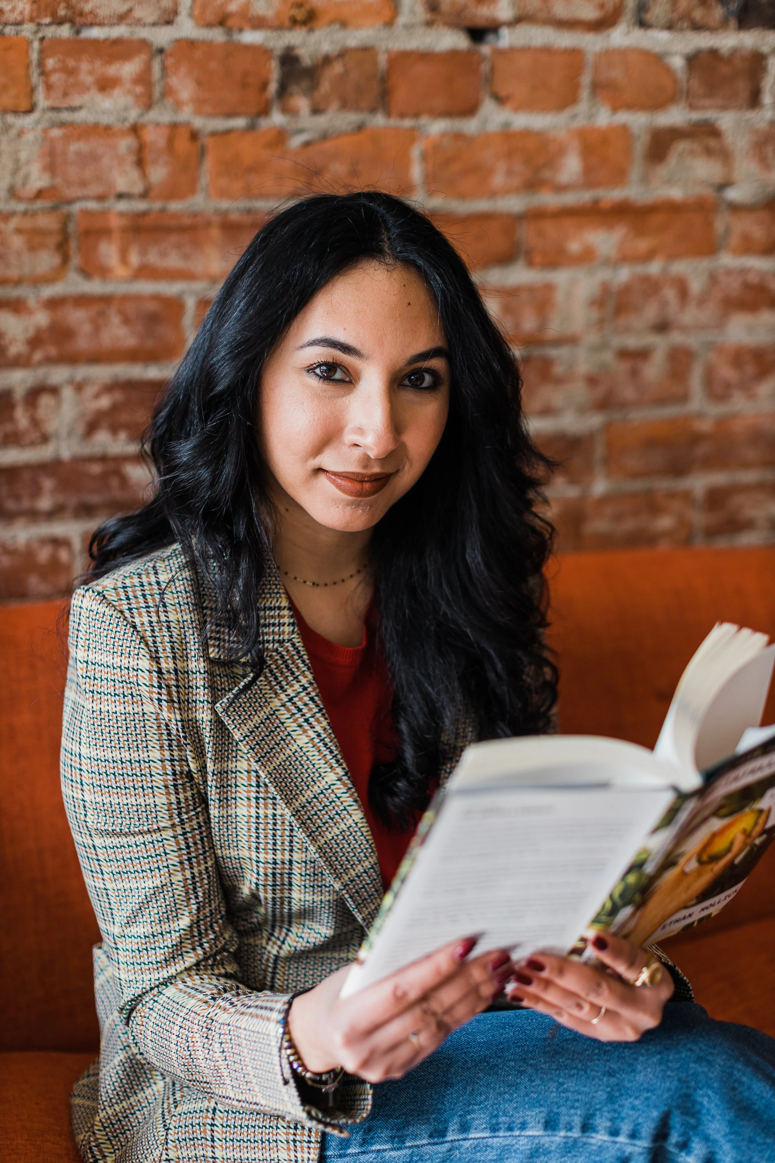A woman with black, wavy hair and tan skin, wearing a plaid blazer and red shirt, reading a magazine in a cozy setting with a brick wall background.