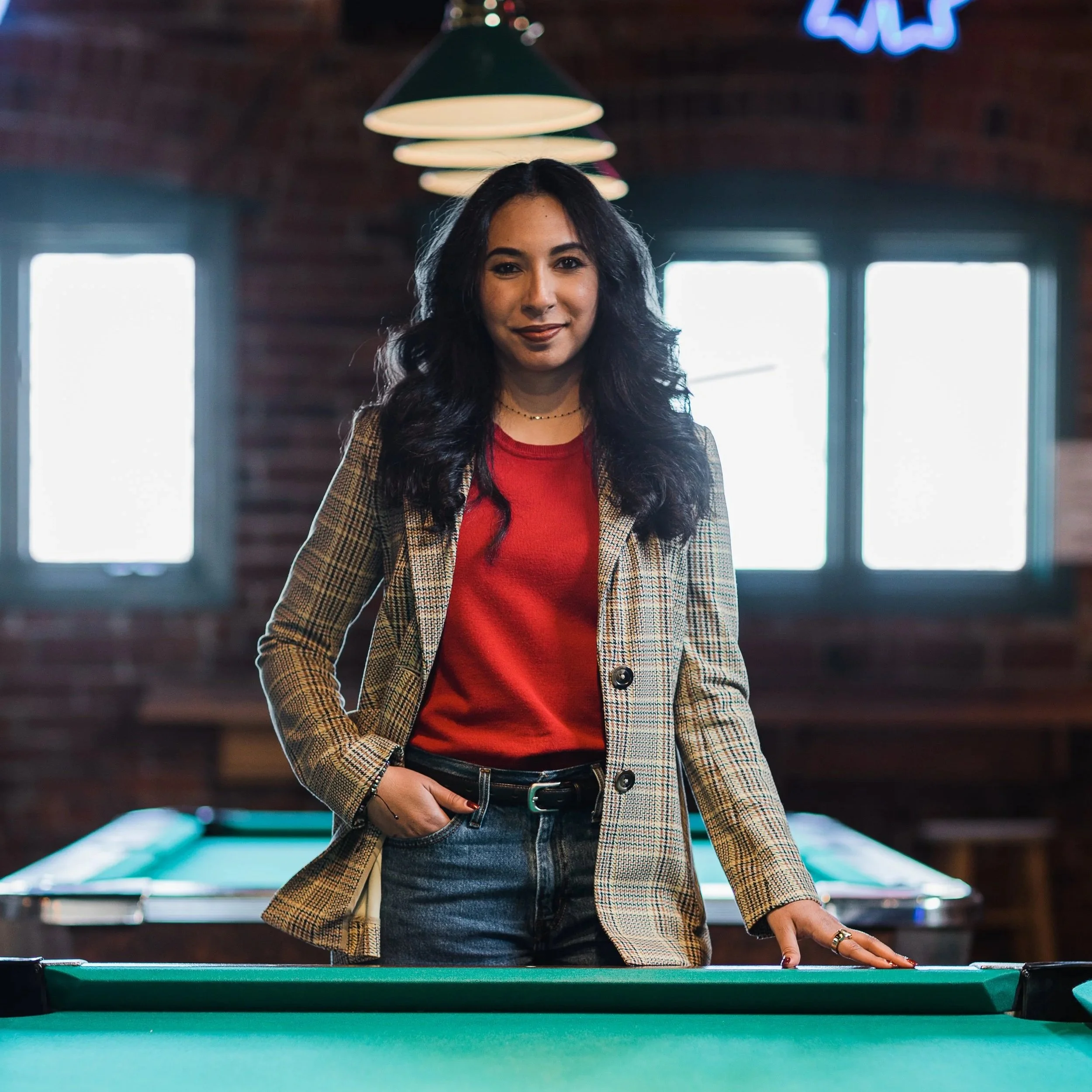 A woman with long dark hair, wearing a red top and plaid blazer, stands by a pool table in a dimly lit room with large windows and neon signs.