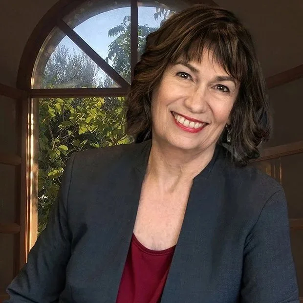 A woman with shoulder-length dark brown hair, smiling, wearing a dark blazer and a maroon top, seated indoors near a large arched window with greenery outside.