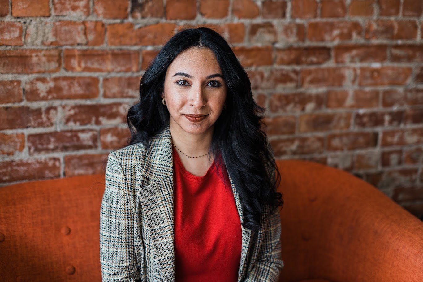 A woman with long black hair and wearing a red top and checked blazer, sitting on an orange couch against a brick wall.
