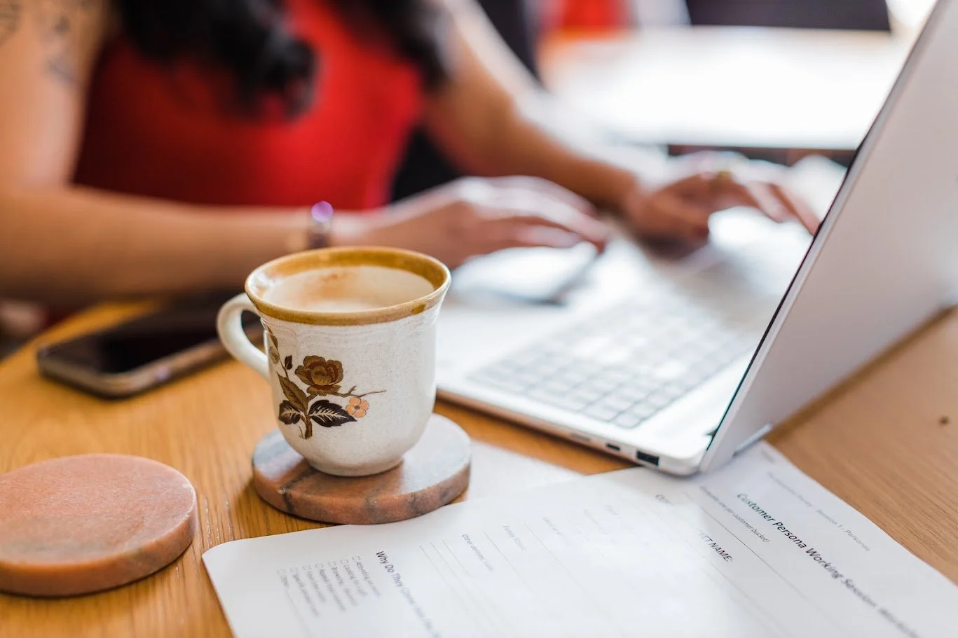 A laptop, a cup of coffee on a coaster, a smartphone, and papers on a wooden table with a person working in the background.