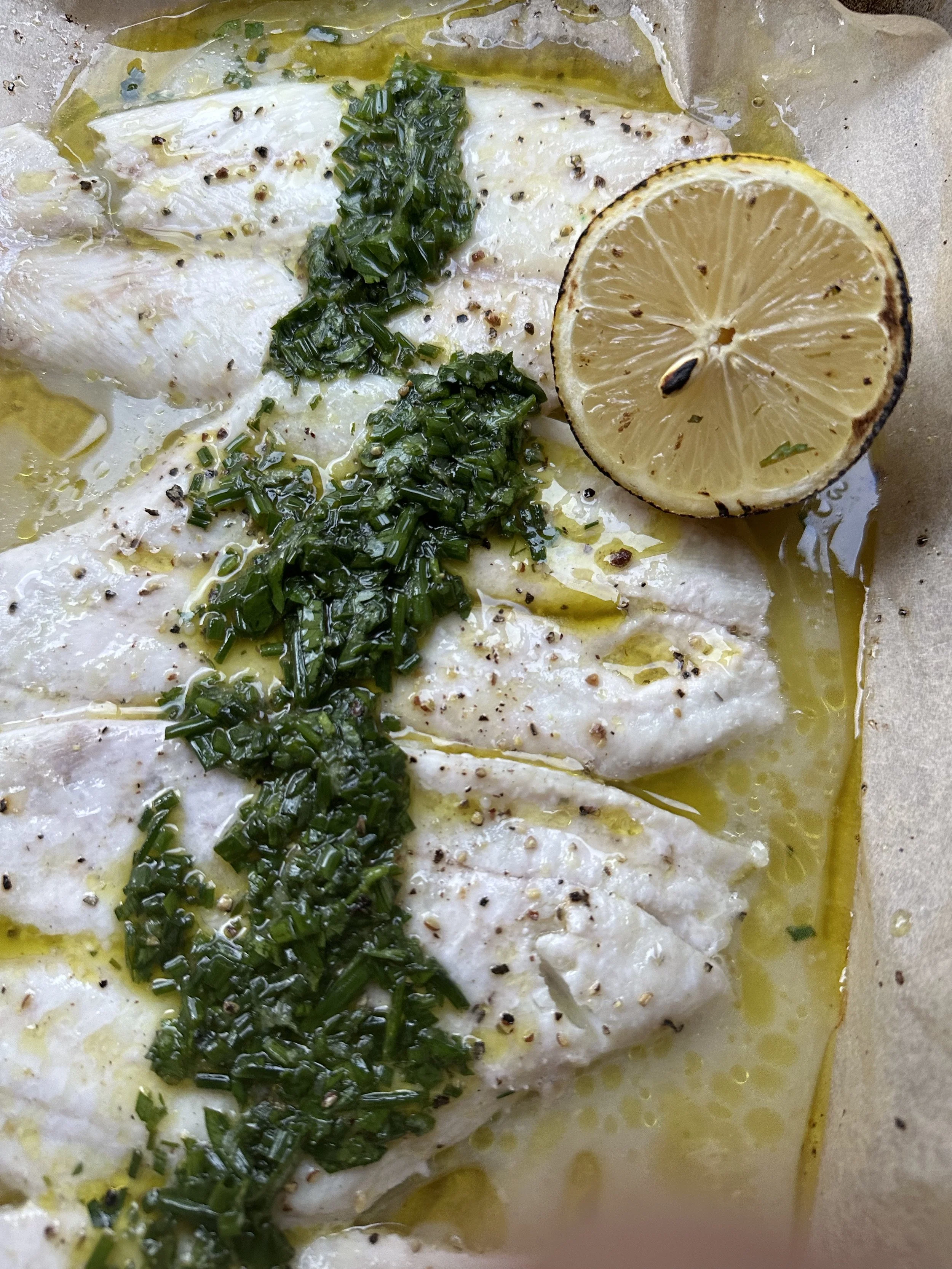 Baked white fish fillet topped with gremolata, sliced lemon, and seasoned with black pepper, baked in a baking dish with lemon juice and olive oil.