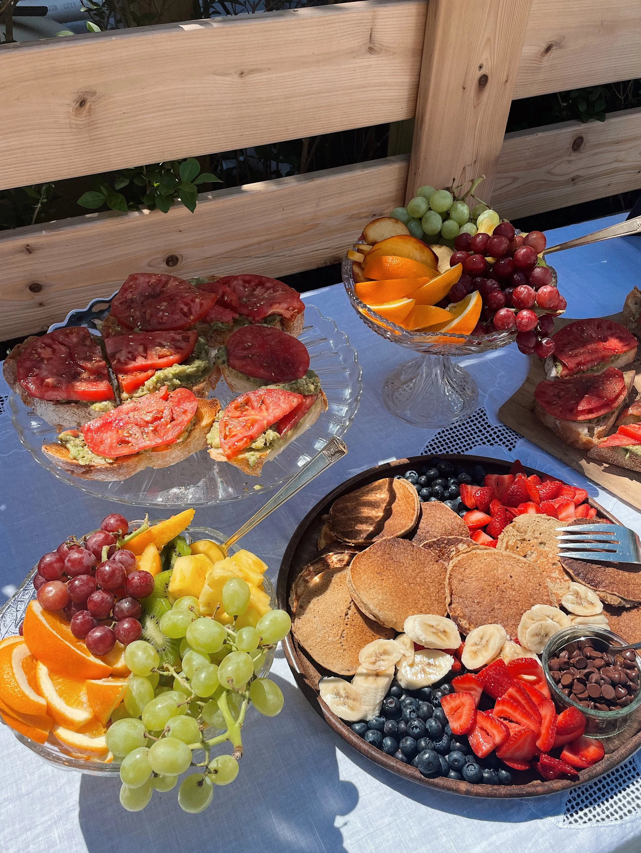 Assorted fruit and breakfast food items on a table with a white tablecloth, including sliced tomatoes on toast, fresh fruit bowls with grapes, oranges, and strawberries, and a platter of pancakes with bananas, blueberries, and chocolate chips.