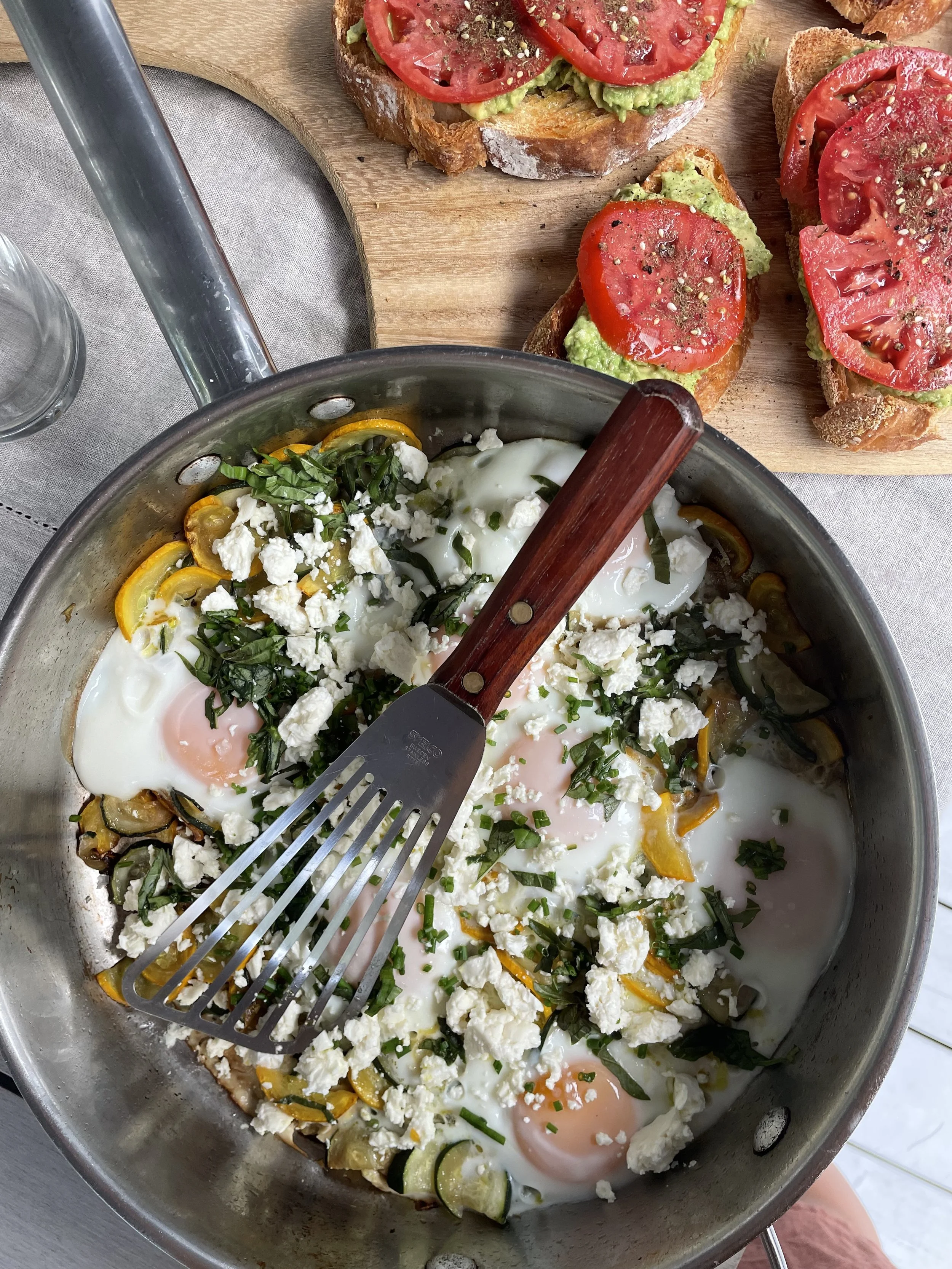 A skillet with baked eggs topped with crumbled cheese, chopped herbs, and sliced vegetables. Nearby on a wooden board are slices of toasted bread topped with avocado and tomato slices, sprinkled with black pepper and herbs.