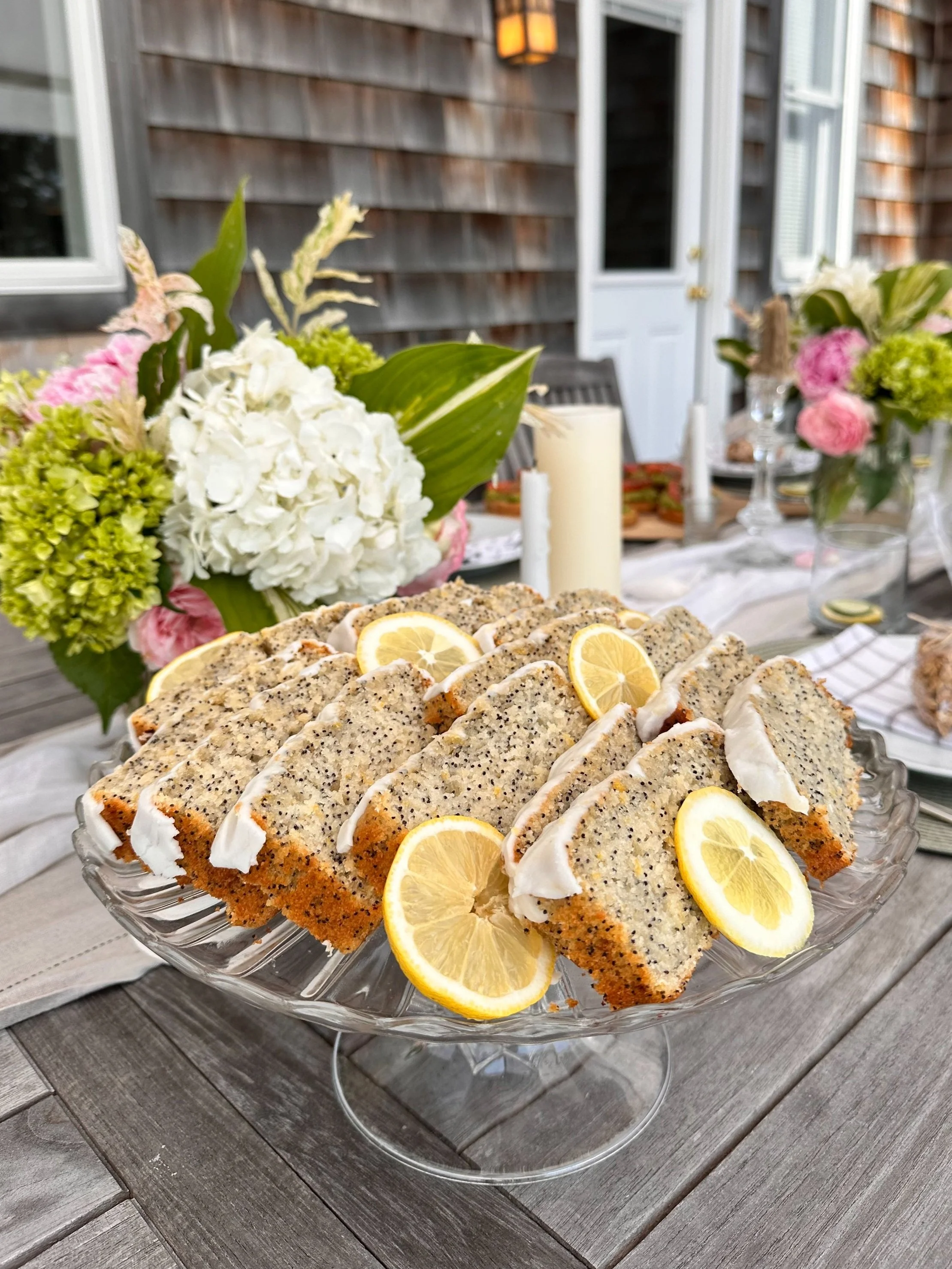 Lemon poppy seed cake slices garnished with lemon slices on a glass cake stand at an outdoor gathering with floral arrangements and candles.