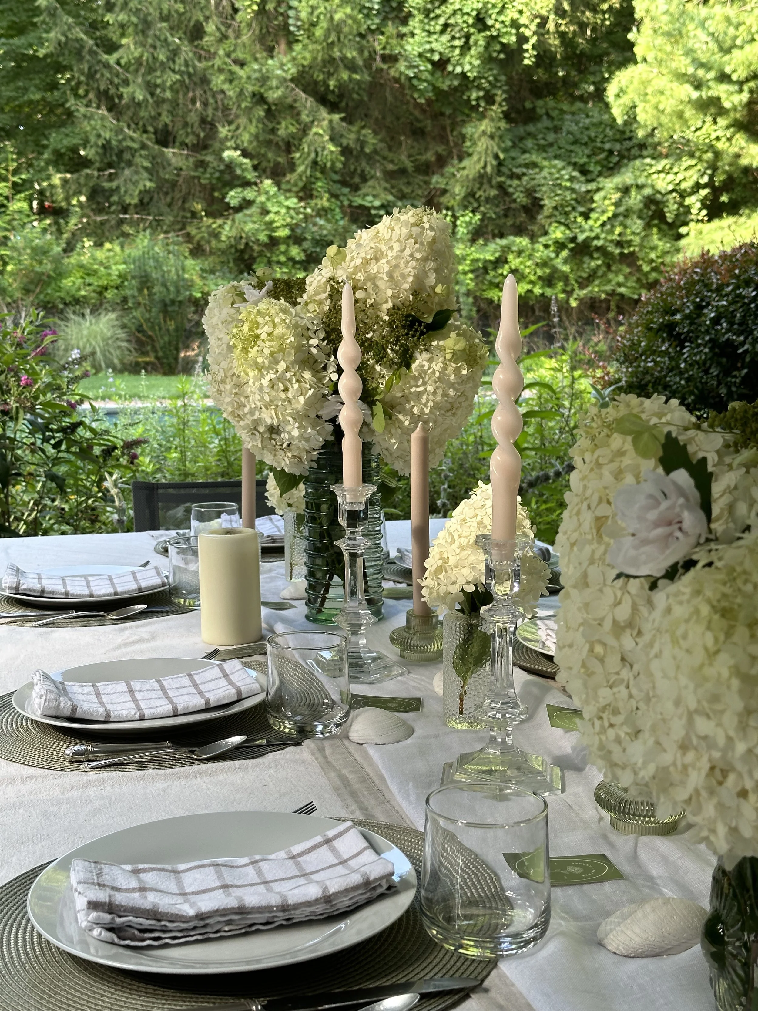 Elegant outdoor dining table decorated with white hydrangea floral centerpieces, pink twisted candles in glass holders, white plates with folded napkins, clear glasses, and seashell accents, set against a lush green garden background.