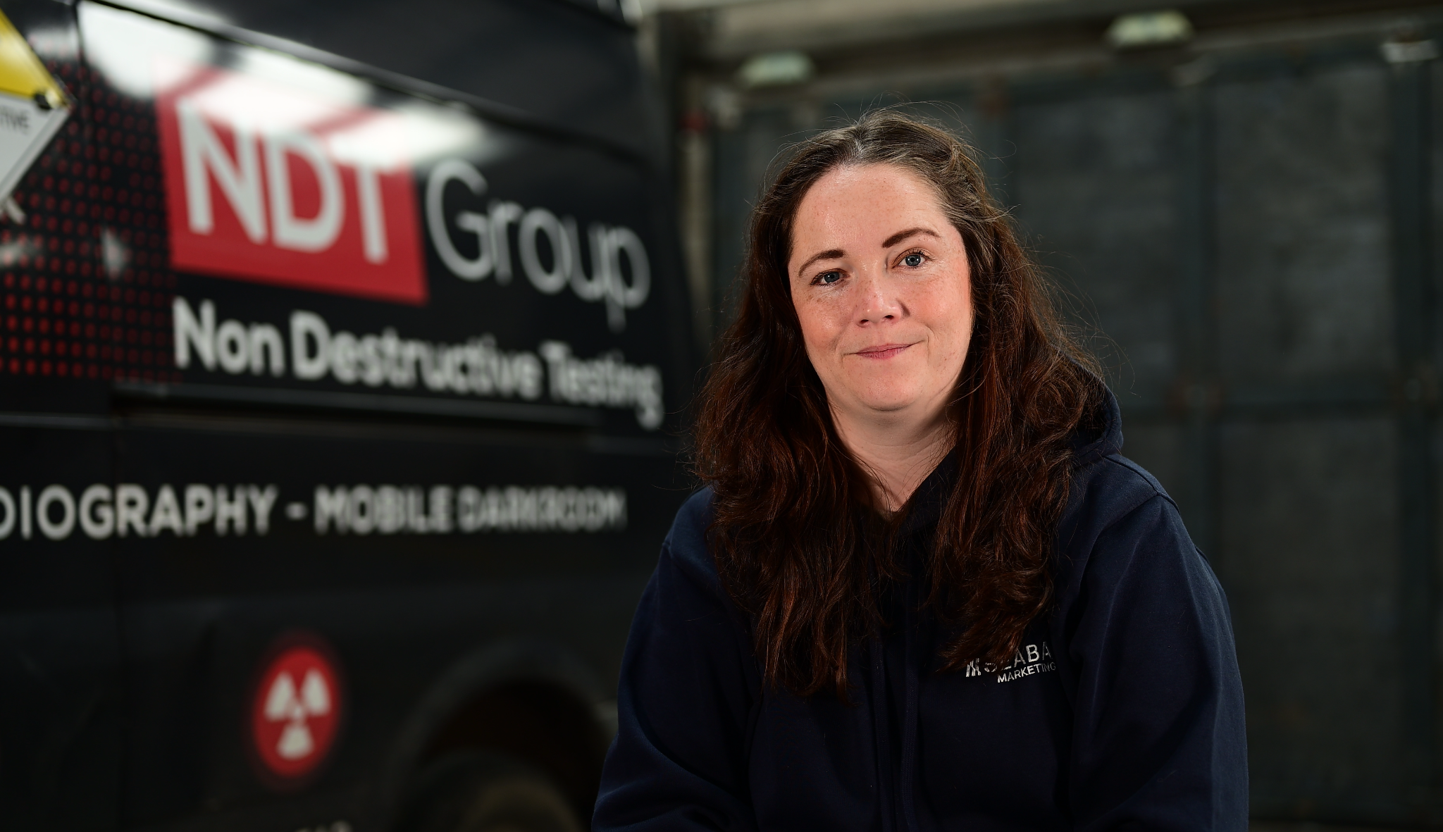 Girl with brown hair, navy jumper sat in front of a black NDT Group van