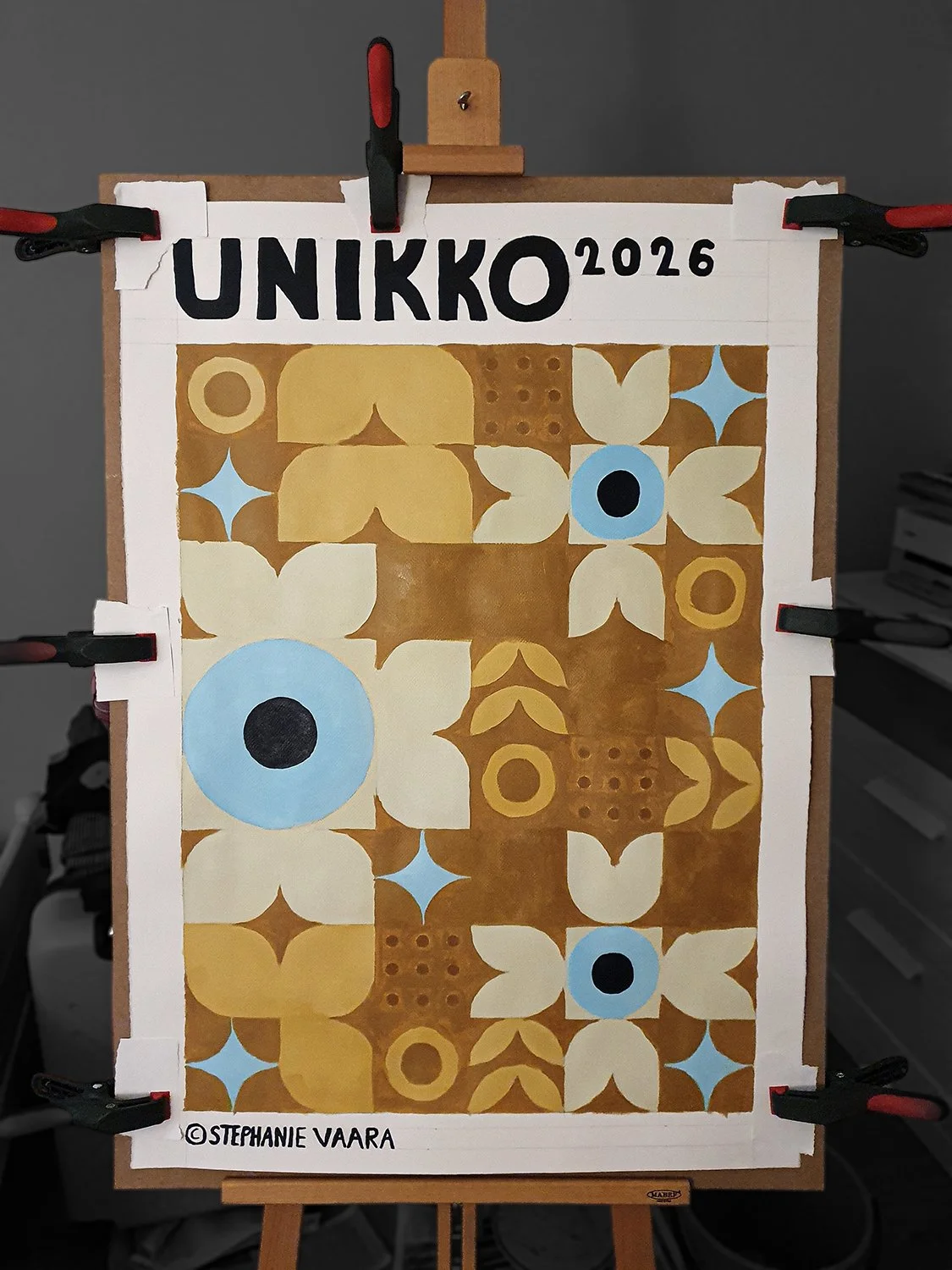 A geometric patterned poster with floral and circle motifs in yellow, blue, white, and brown tones, mounted on an easel with black and red clamps, titled 'UNIKKO 2026' at the top, signed by Stephanie Vaara at the bottom.