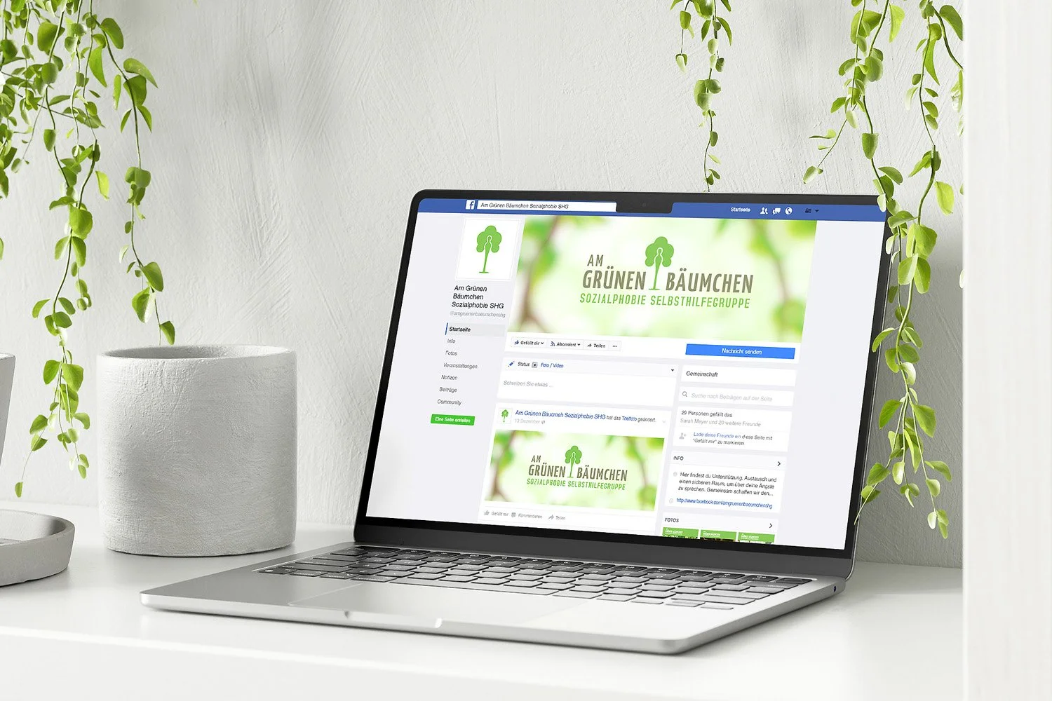 A laptop on a white desk displays a Facebook page for a group called 'Am Grünen Bäumchen'. The page has a green and white logo with a tree icon, and the cover photo features a blurred green background with text in German. Surrounding the laptop are t