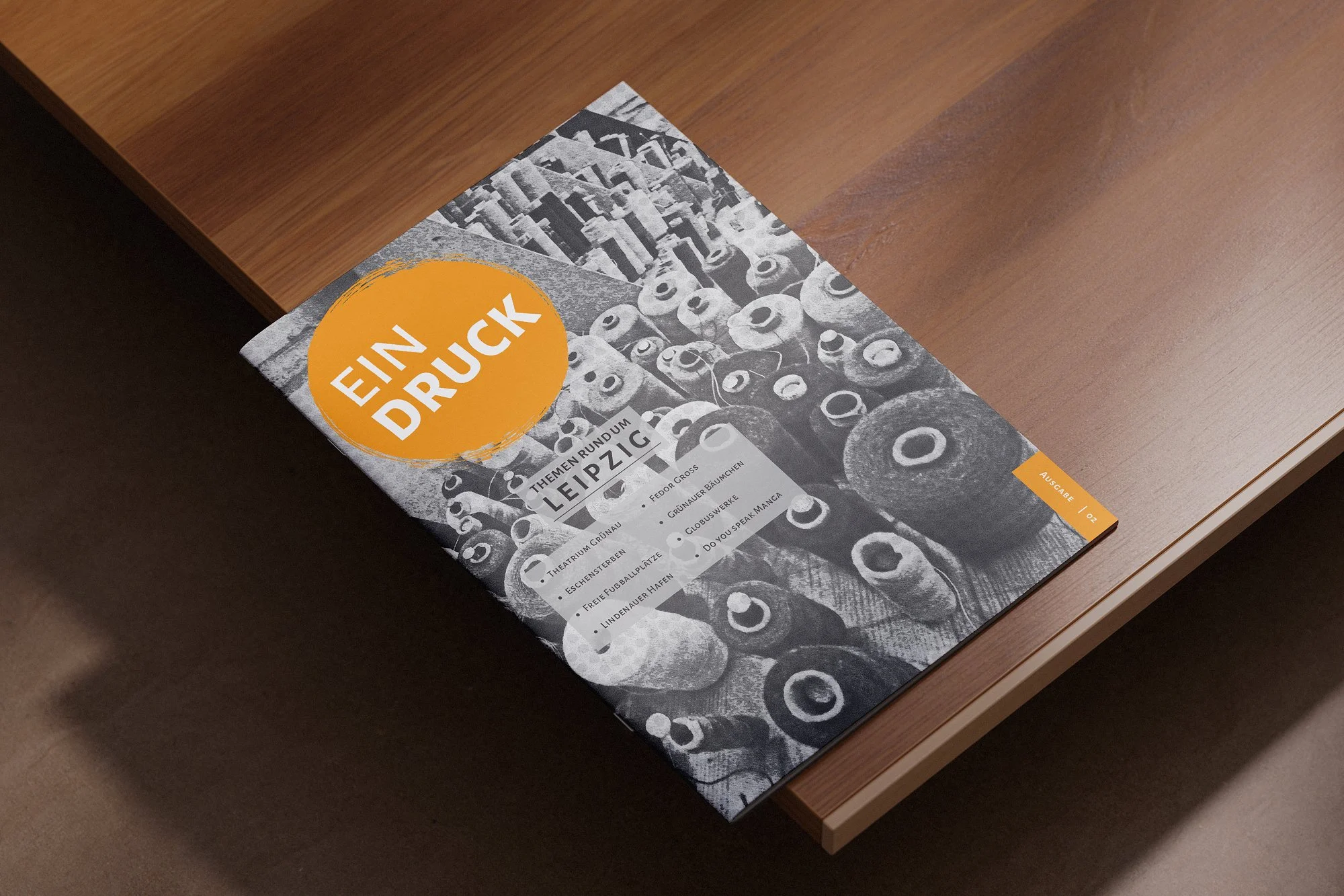 A magazine lying on a wooden table with a black and white cover featuring images of pipes and a large orange circle with white text that reads 'EIN DRUCK'. The magazine title mentions themes around Leipzig and local landmarks.