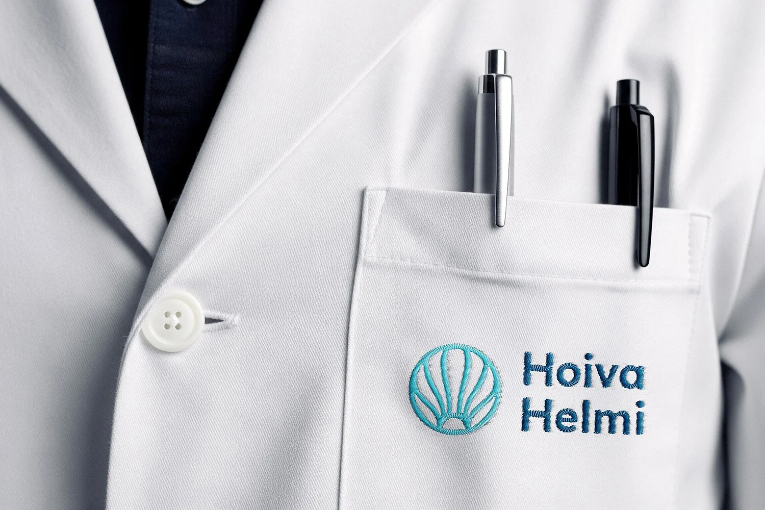 Close-up of a white lab coat with the logo and name 'Hoiva Helmi' embroidered on the chest pocket, featuring two pens tucked in the pocket.