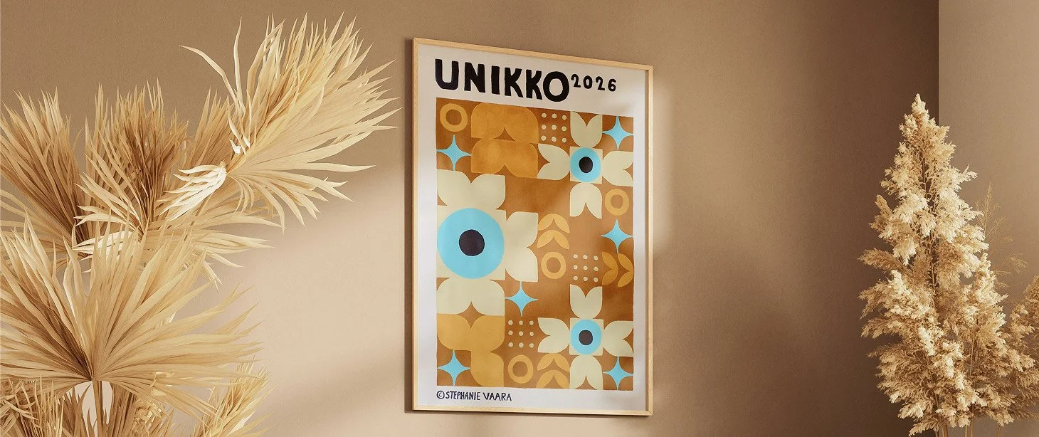 Decorative framed poster with geometric floral patterns in blue, white, gold, and brown colors, labeled "UNIKKO 2026" by Stephanie Vaara, hanging on a beige wall, with dried pampas grass on the left and a dried floral arrangement on the right.
