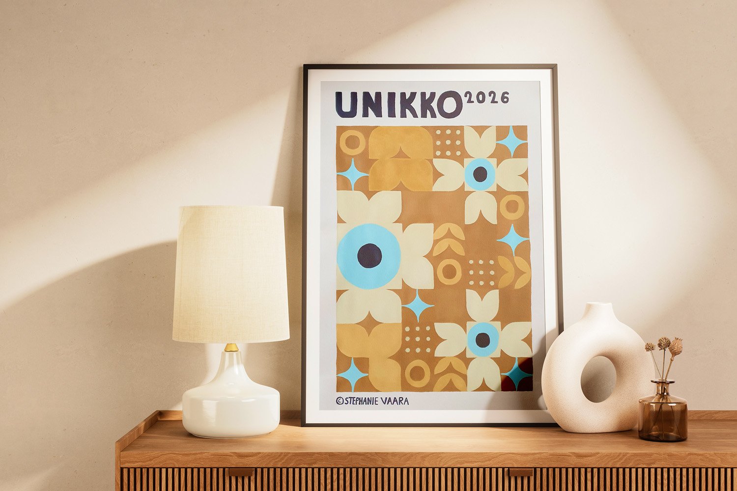 Decorative table with framed artwork titled 'UNIKKO 2026' featuring a floral pattern in beige, blue, and brown, accompanied by a beige lamp, a white ceramic sculpture with a circular hole, and a small amber glass vase with dried flowers.