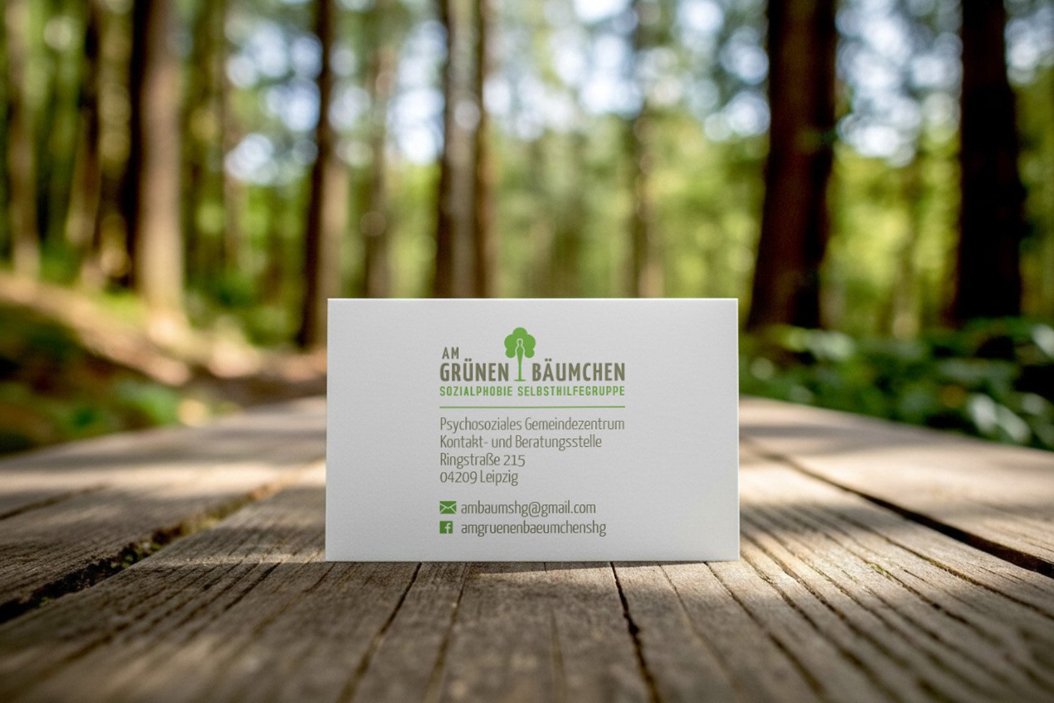 A business card for 'Am Grünen Bäumchen', a social self-help group, placed on a wooden surface outdoors with a forest background.