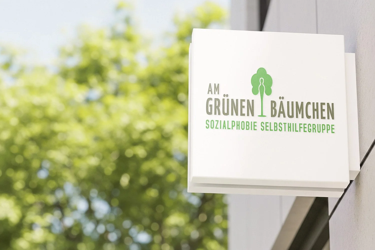 A white signboard outdoors with green text and a tree logo, reading in German 'Am Grünen Bäumchen' and 'Sozialphobie Selbsthilfegruppe', against a background of blurred green trees.