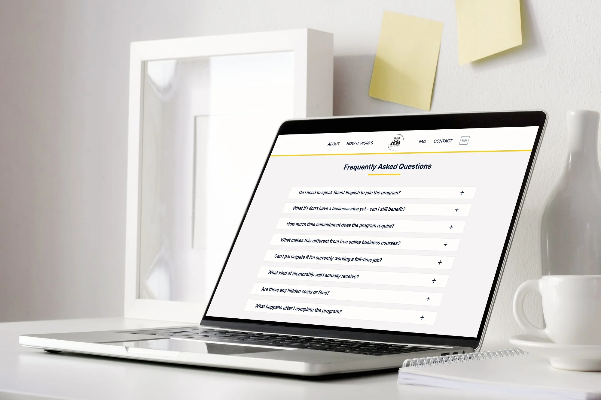 Laptop on a white desk displaying a FAQ page about online business programs, with a white cup, a notebook, and a vase nearby, in front of a white wall with sticky notes.