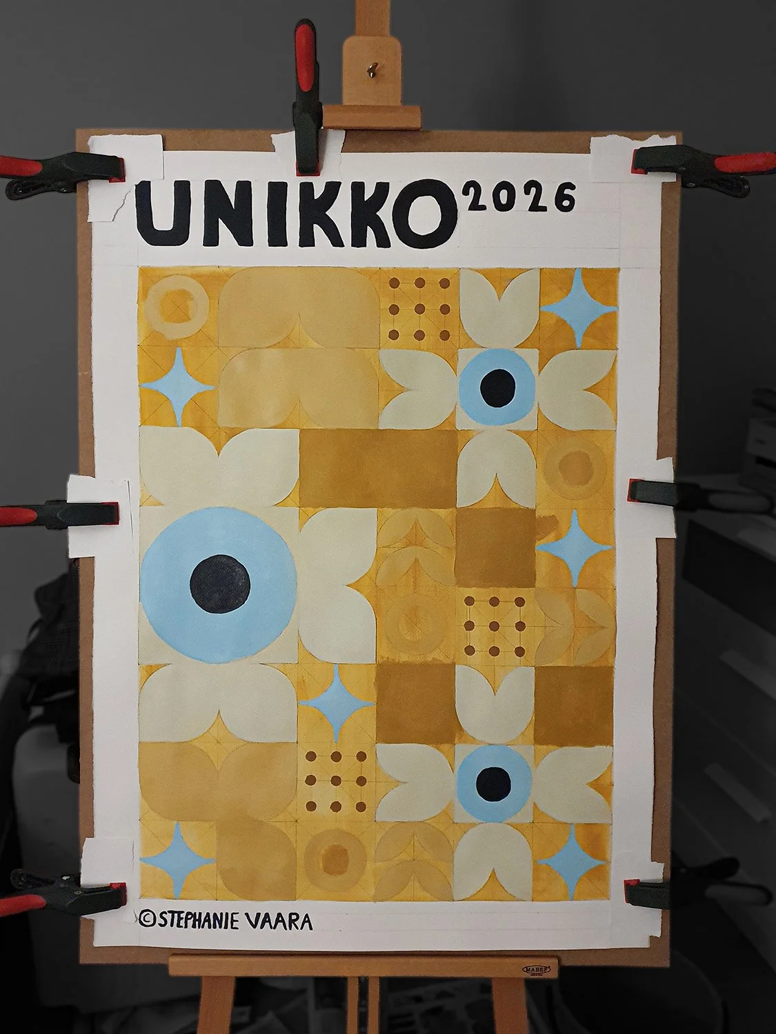 Abstract geometric art with yellow, blue, and beige shapes on a board, titled UNIKKO 2026 by Stephanie Vaara, secured with clamps on an easel.