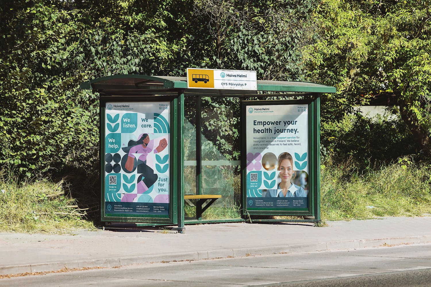 A bus stop shelter with advertising posters on the sides, surrounded by greenery and trees, with a paved sidewalk in front.